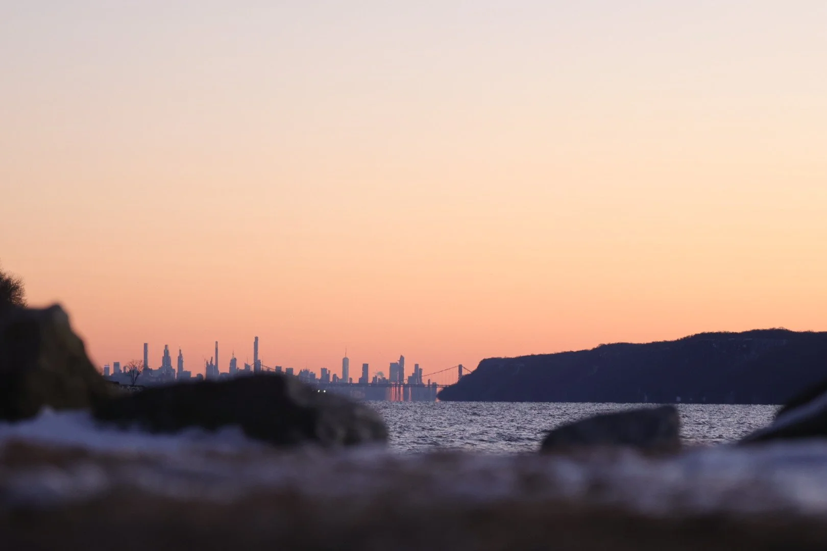 City skyline at sunset viewed from across a body of water with rocks in the foreground and hills on the right.
