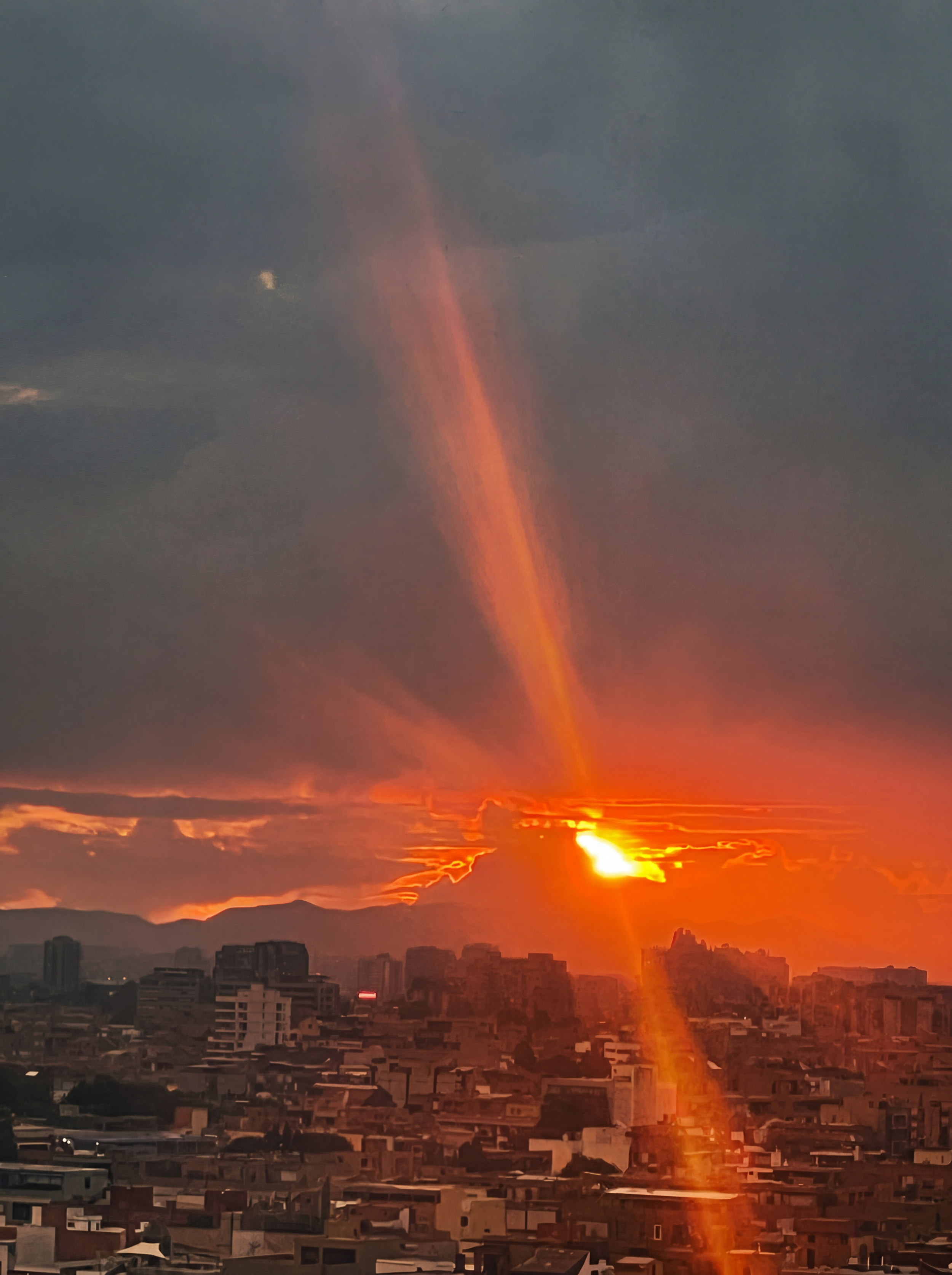 Cityscape during sunset with the sun partially below the horizon, casting an orange glow over buildings and mountains. A vivid beam of sunlight stretches vertically in the sky, illuminating clouds and creating a dramatic atmosphere.