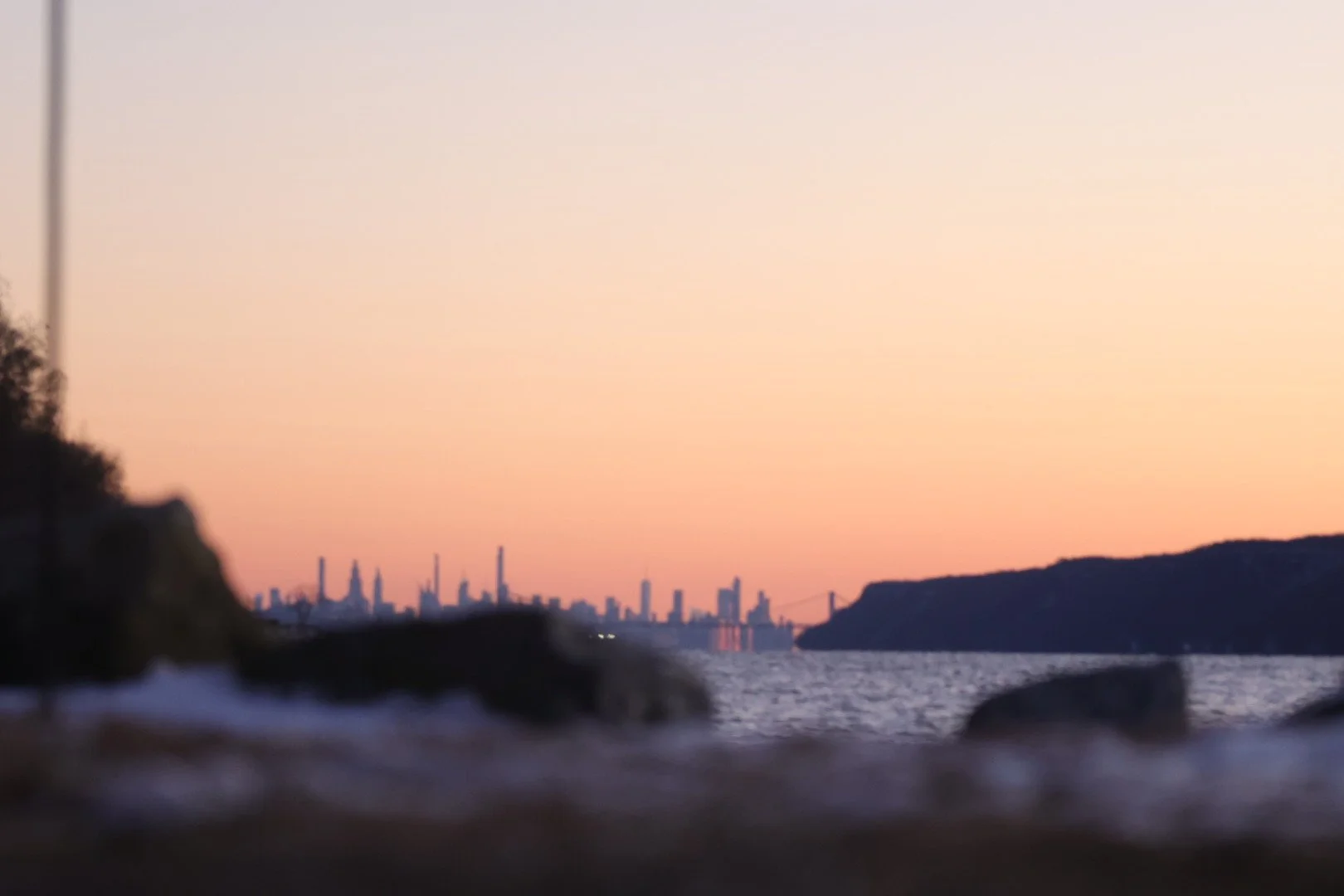 City skyline at sunset viewed from a rocky shoreline with water in the foreground.
