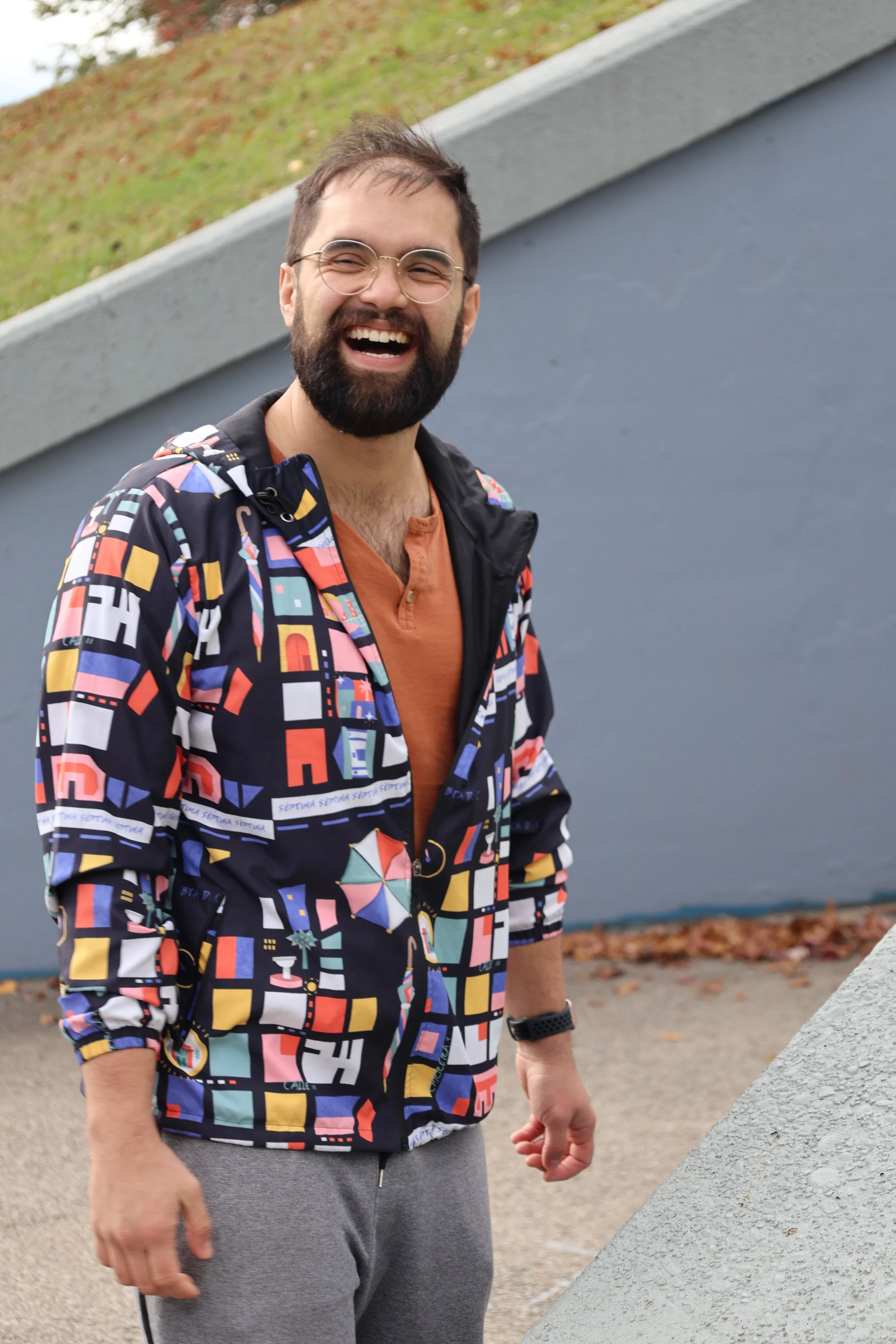 A smiling man with glasses and a beard wearing a colorful geometric-patterned jacket, a burnt orange shirt, gray sweatpants, and a black watch, standing outdoors near a concrete structure, with grass and fallen leaves in the background.
