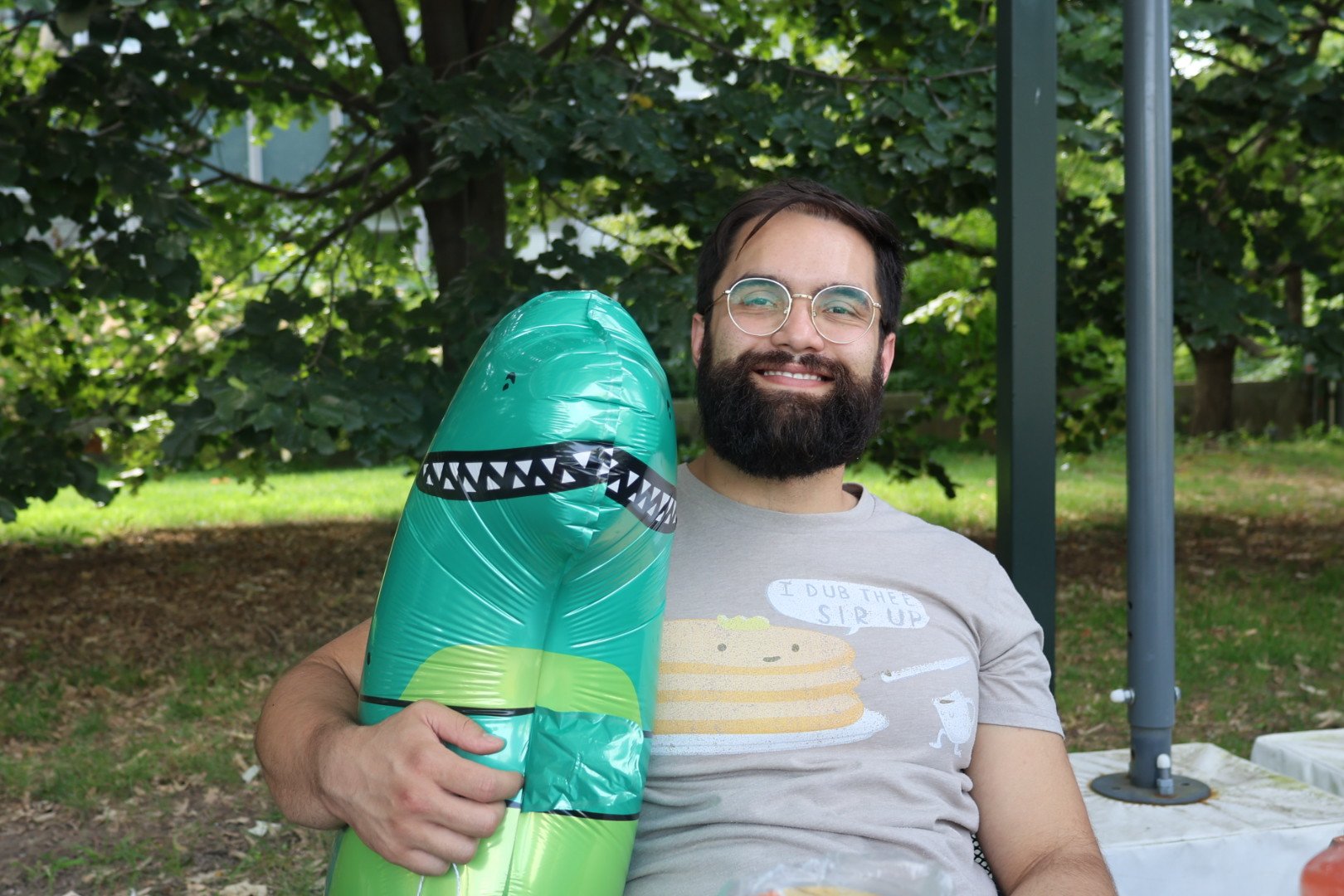 A smiling man with glasses and a beard sitting outdoors in a park, holding a green inflatable shark toy. He is wearing a light gray t-shirt with a pancake and syrup illustration and a speech bubble that says 'I DUB THEE SIR UP.' Background has green trees and grass.