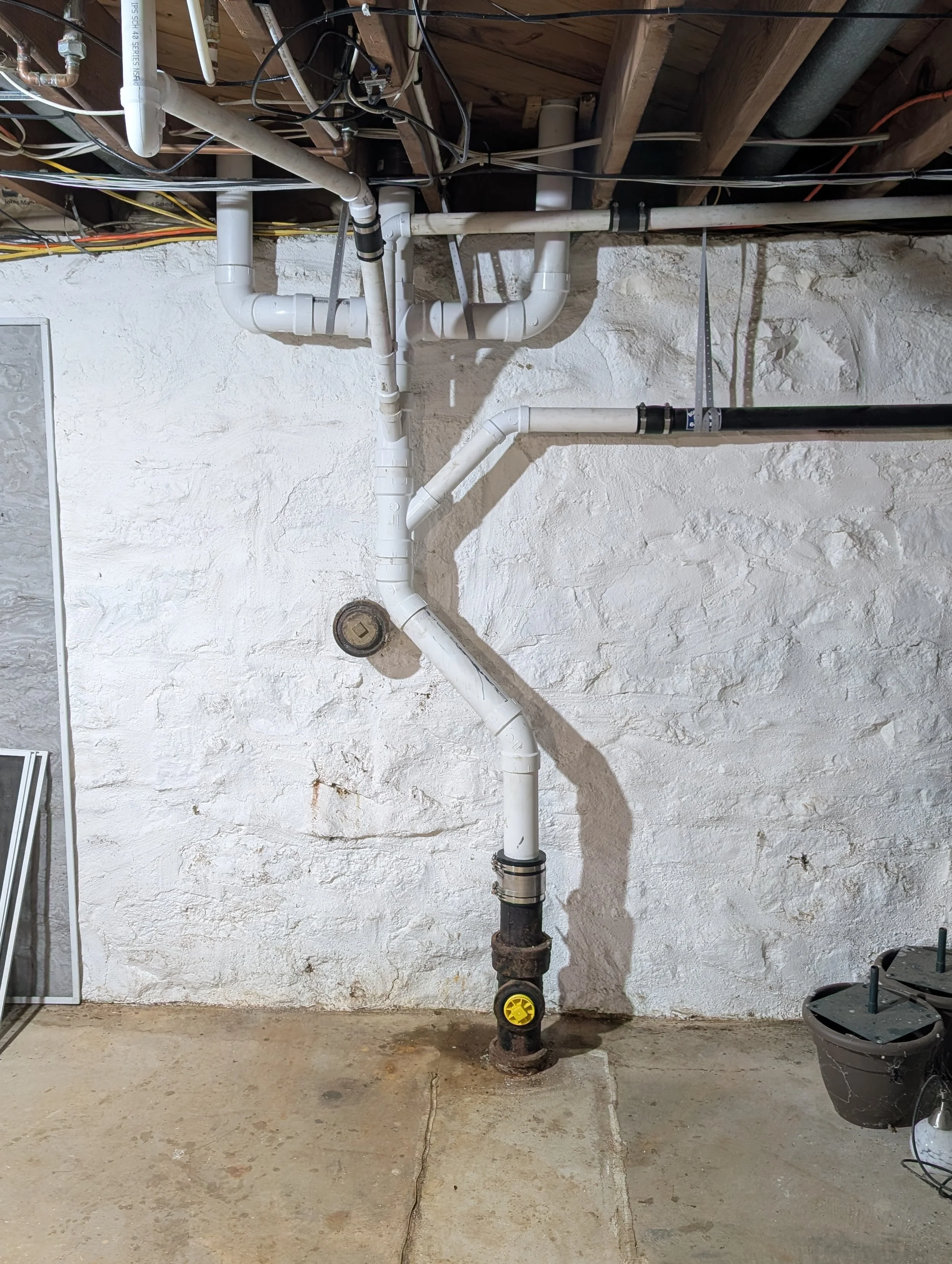 Plumbing pipes and fittings installed in a basement or utility room, with a concrete floor and a textured wall, and some electrical wiring overhead.