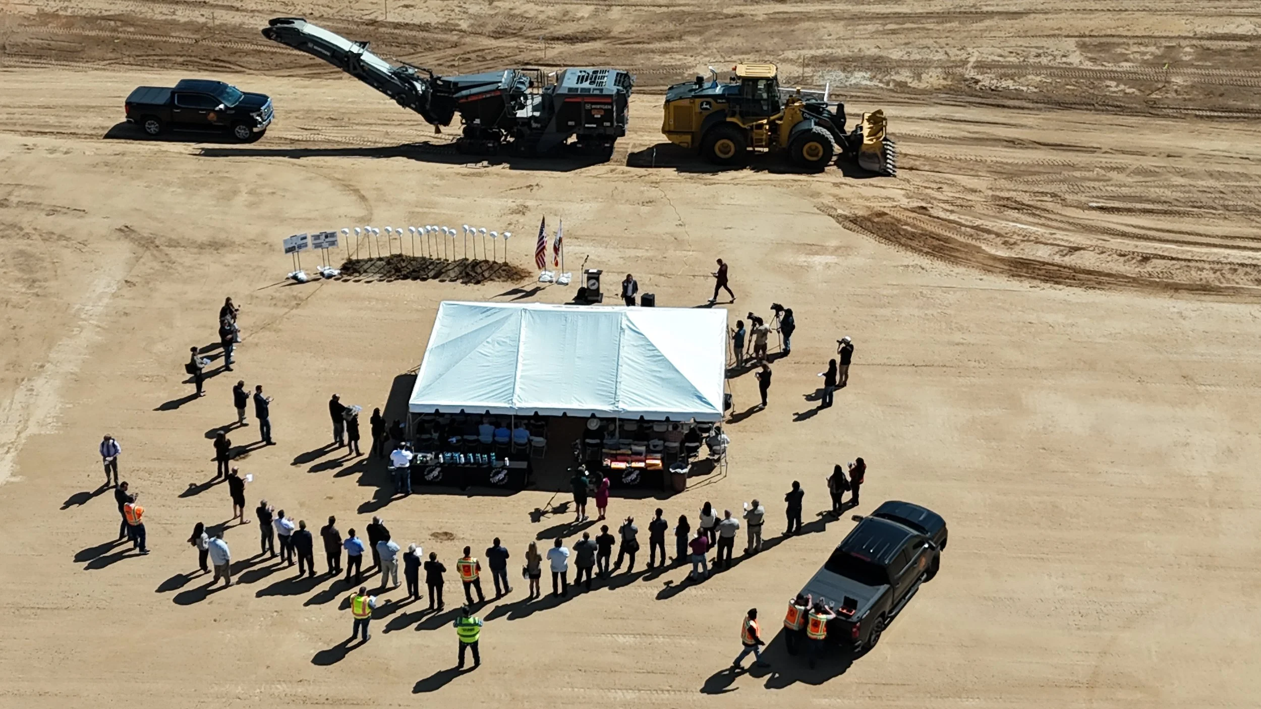 An aerial view of the groundbreaking site. A white tent in the center with people standing in a semi-circle on the south side. The groundbreaking shovels and helmets on top are on the north side. Further up is the black asphalt grinder machine.