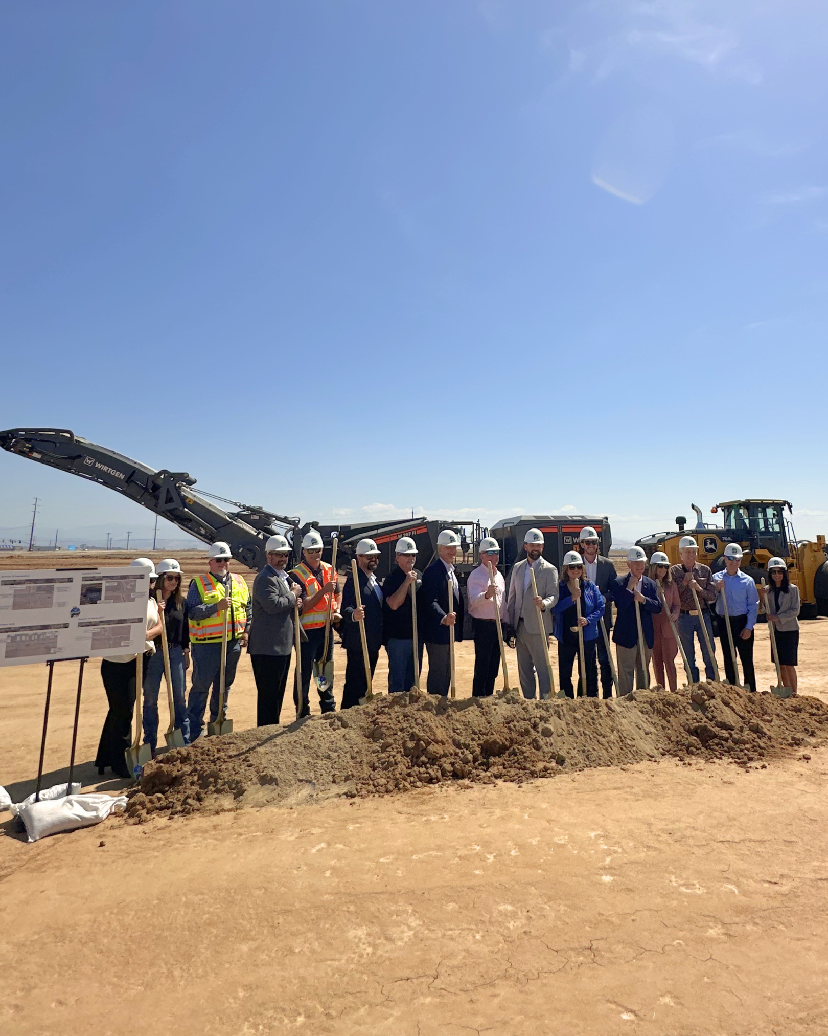 All groundbreaking participants posing behind golden shovels in a mound of dirt and they're wearing hard hats with the new State Route 41 logo. A black asphalt grinder machine behind all of them.