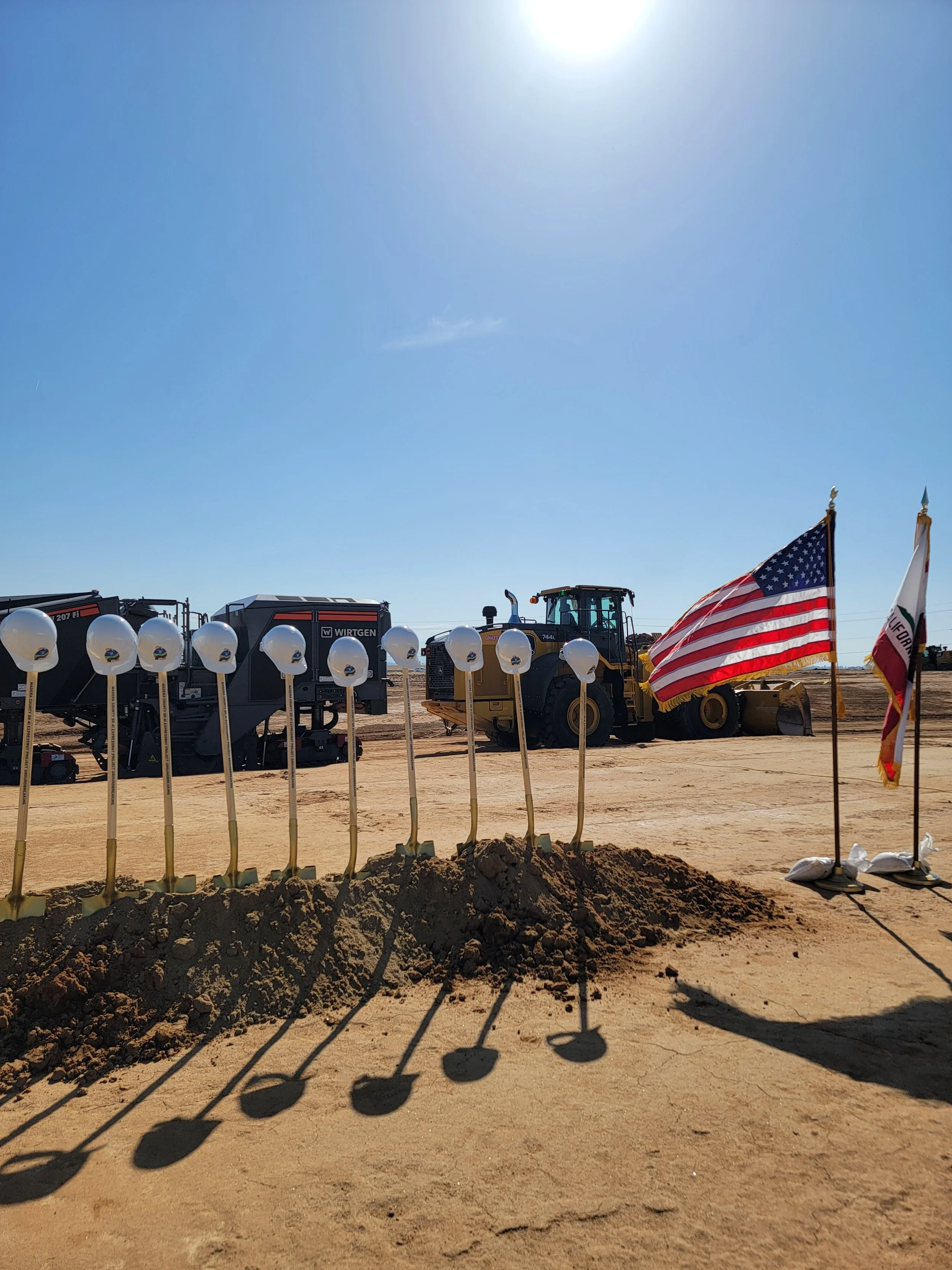 Mound of dirt with the inserted golden shovels and white hard hats on top going from the left side to 3/4 of the image. The United States and California state flag are on the right of the shovels. A black asphalt grinder behind everything.
