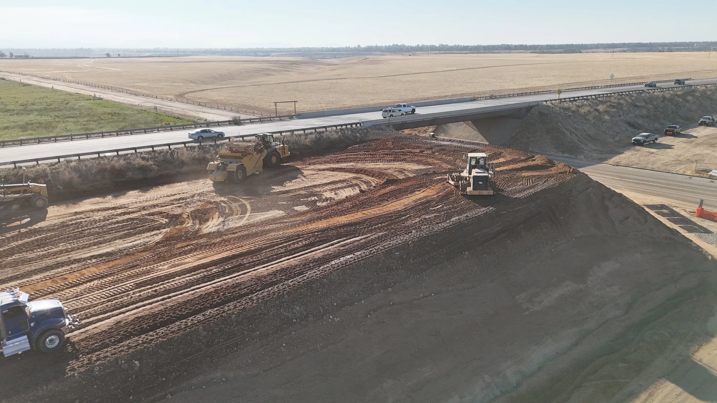 A scraper, motor grader, and compactor work on the soil besides Avenue 11 bridge.