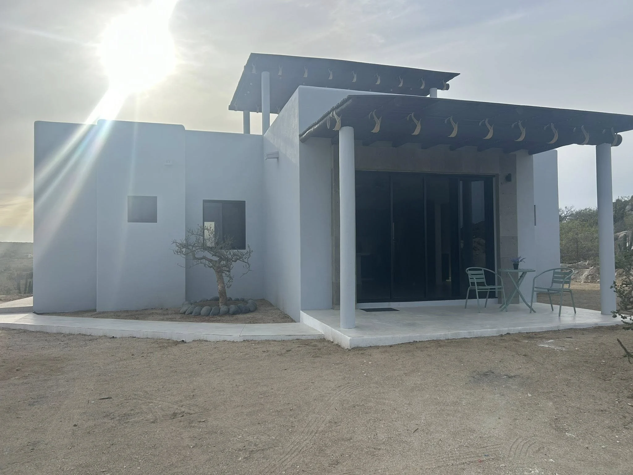 Exterior view of a white modern house with a flat roof, patio furniture, and a small tree, under a bright sky with sunlight.