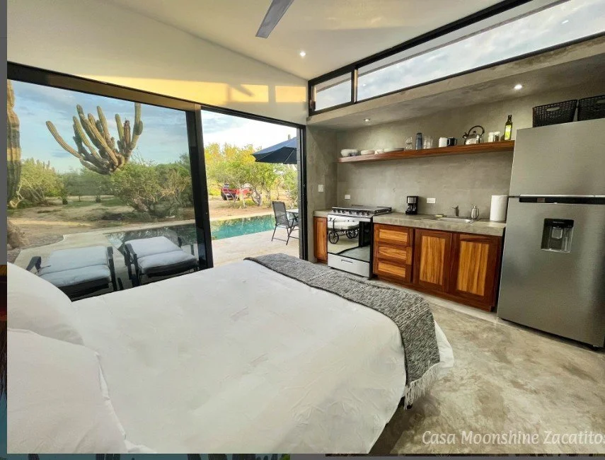 A modern studio with a bed near large glass sliding doors leading to a patio and backyard with desert plants and a cactus. The room includes a small kitchen with a refrigerator, stove, sink, and open shelving. Outside, there is patio furniture and a swimming pool.