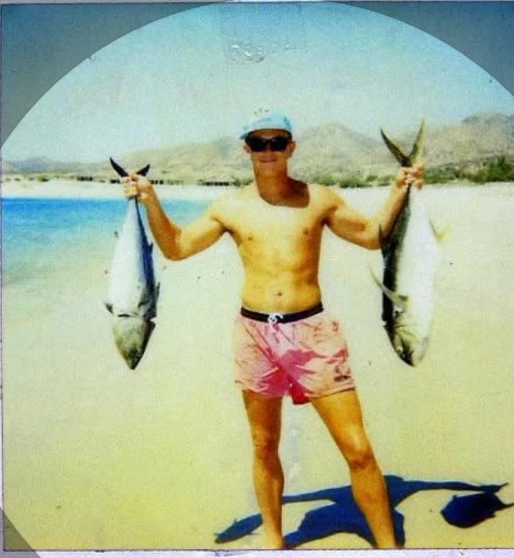 A man wearing pink swim trunks, sunglasses, and a cap, holding two fish on a beach with mountains and water in the background.
