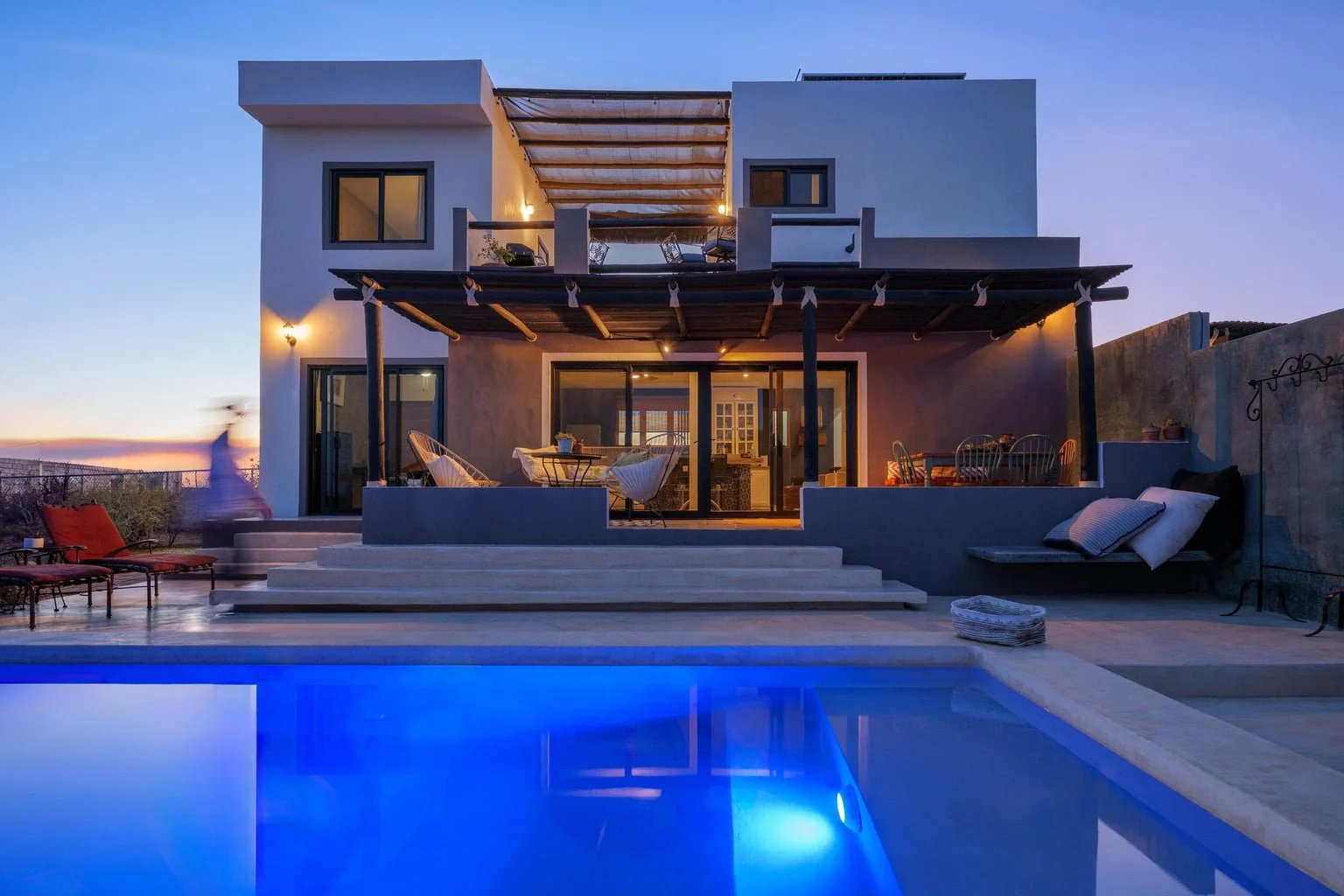 Modern two-story house with large sliding glass doors, a pool in the foreground, outdoor seating area, and sunset sky in the background.