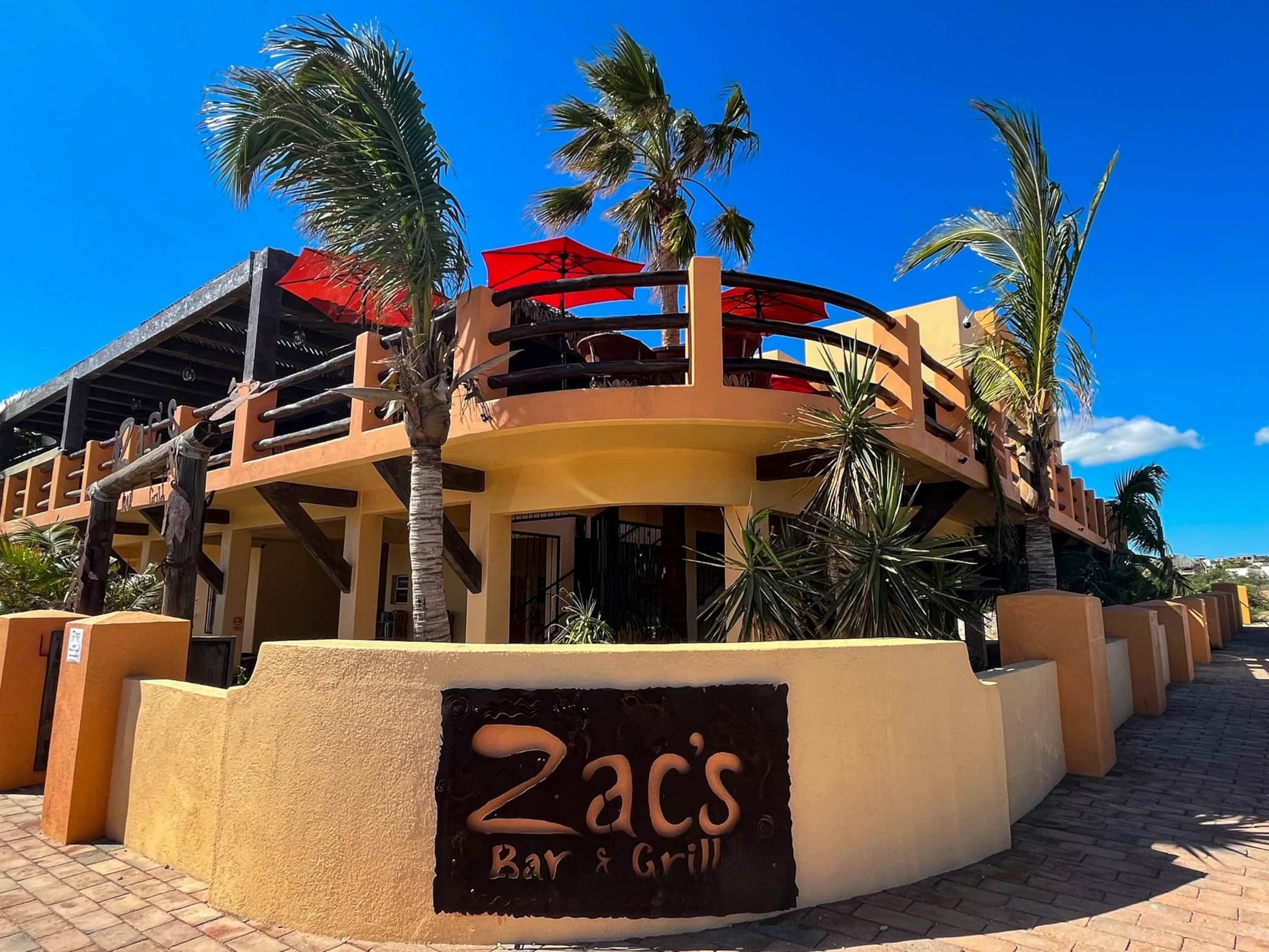 Exterior view of a Mexican restaurant called Zacs Bar & Grill with a yellow stucco wall, a decorative metal sign, palm trees, and red umbrellas on a balcony above.
