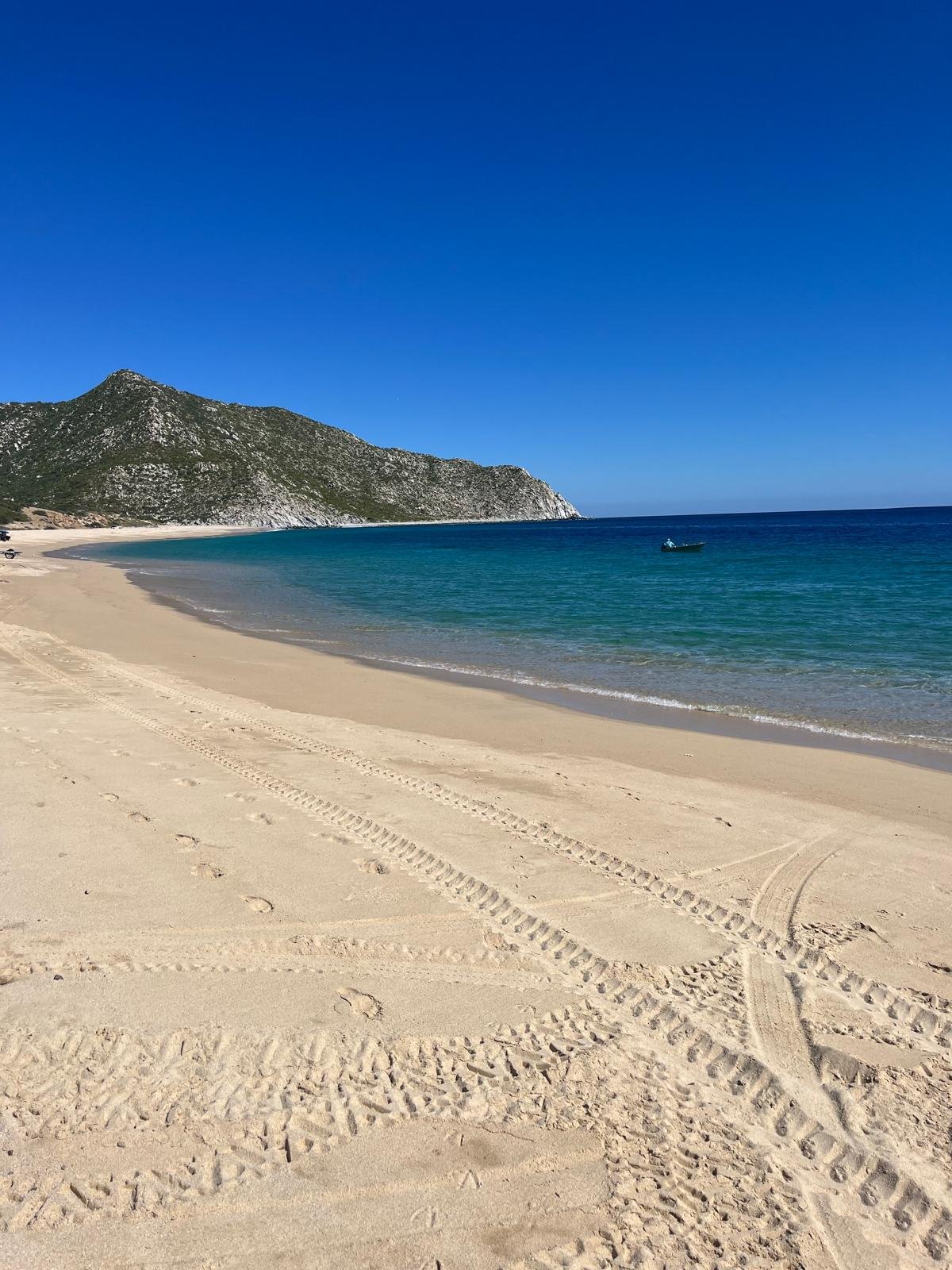 A sandy beach with tire tracks and footprints, facing calm azure water with a small boat, bordered by green hillside under a clear blue sky.