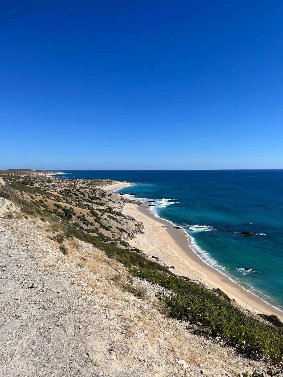 Scenic view of a beach with sandy shoreline, rocky outcrops, and blue ocean under a clear blue sky, seen from a hillside.