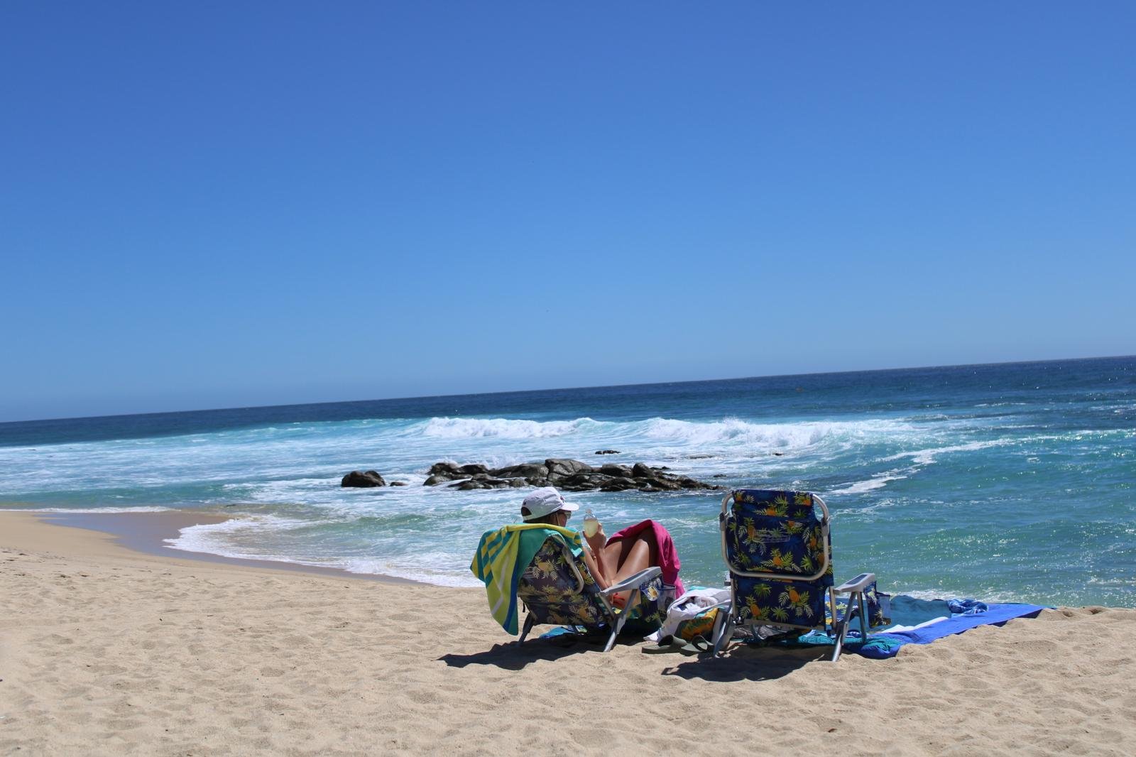 People relaxing on a sandy beach with a clear blue sky and ocean waves, sitting in beach chairs with towels and belongings nearby.
