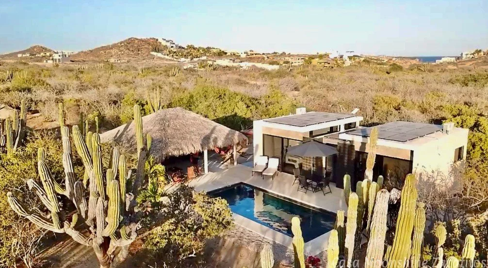 Aerial view of a desert house with a swimming pool, surrounded by tall cacti and desert vegetation, with hills and mountains in the background.