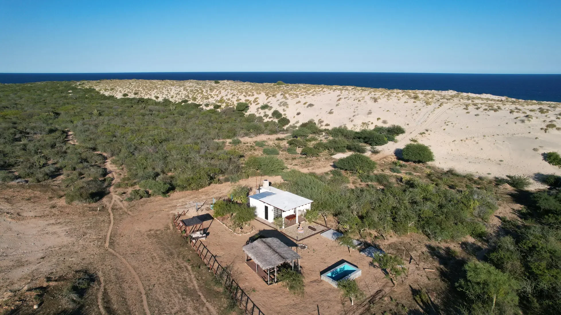 An aerial view of a house with a swimming pool surrounded by greenery and sand dunes, extending to the blue ocean under a clear sky.