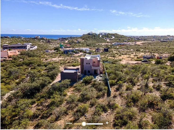 Aerial view of a developing residential area in a desert landscape with sparse vegetation, modern houses, and a distant coastline with the ocean under a partly cloudy sky.
