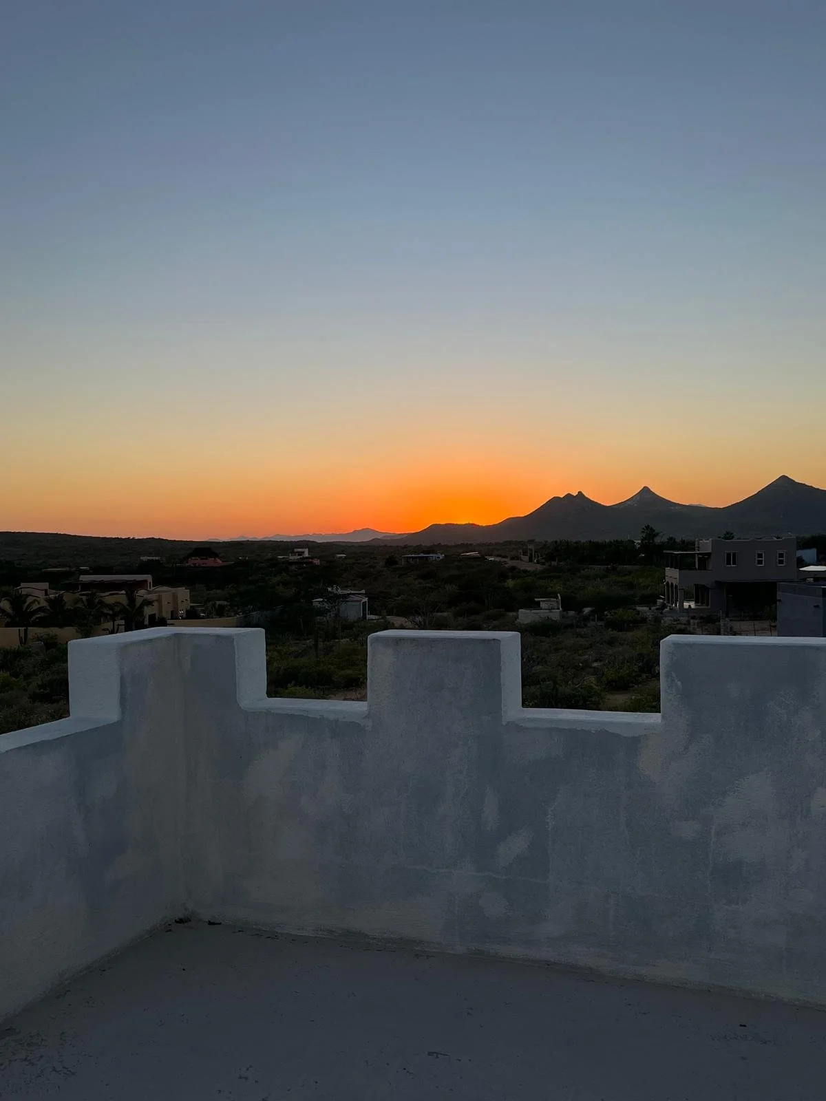 Sunset over mountains with a clear sky, viewed from a rooftop terrace with a white concrete parapet wall.