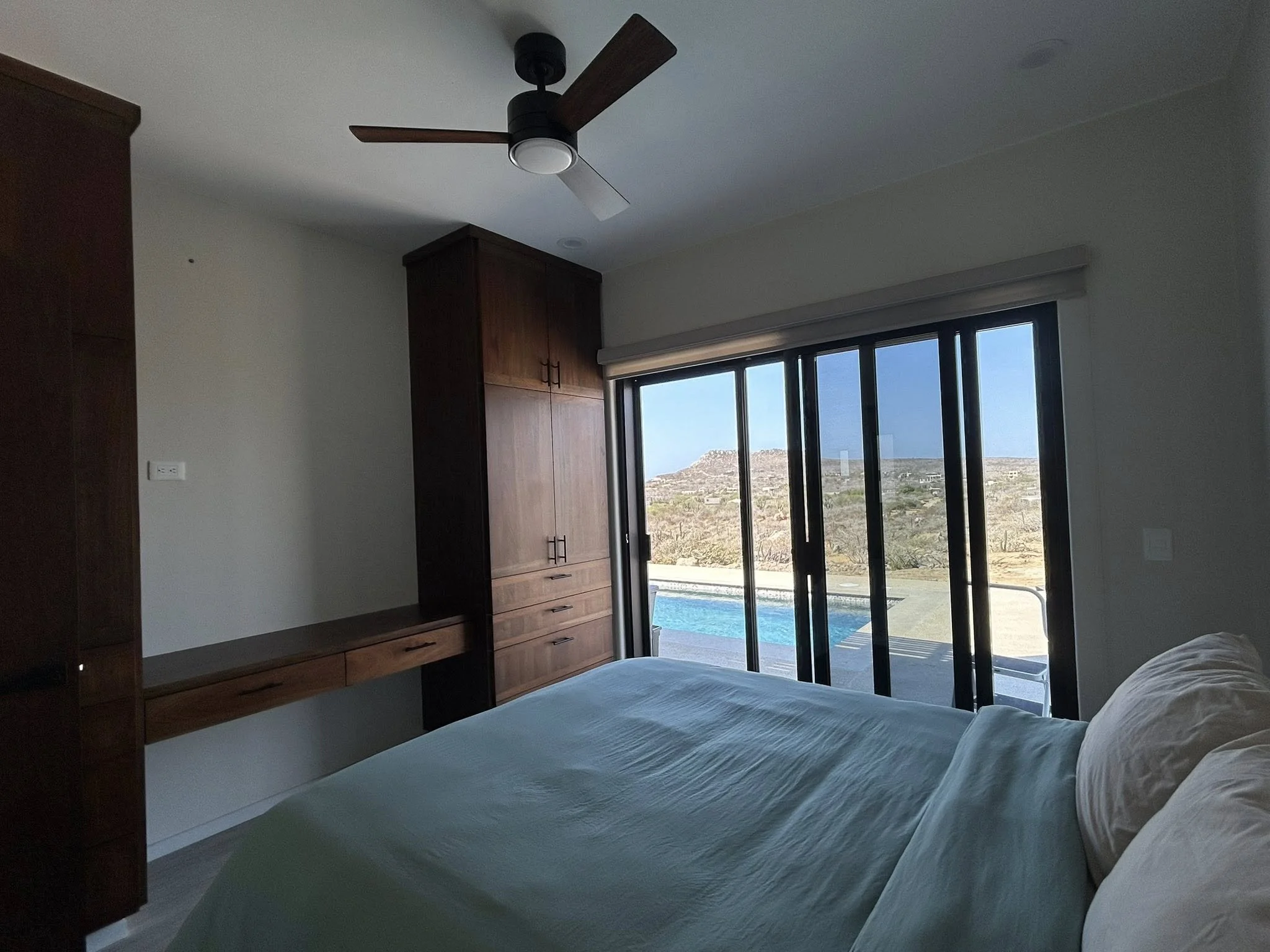 A bedroom with a large bed covered in white linens and pillows, a wooden wardrobe on the left, a sliding glass door with a view of mountains and desert outside, a ceiling fan with wooden blades, and a flat-screen TV mounted on the wall.