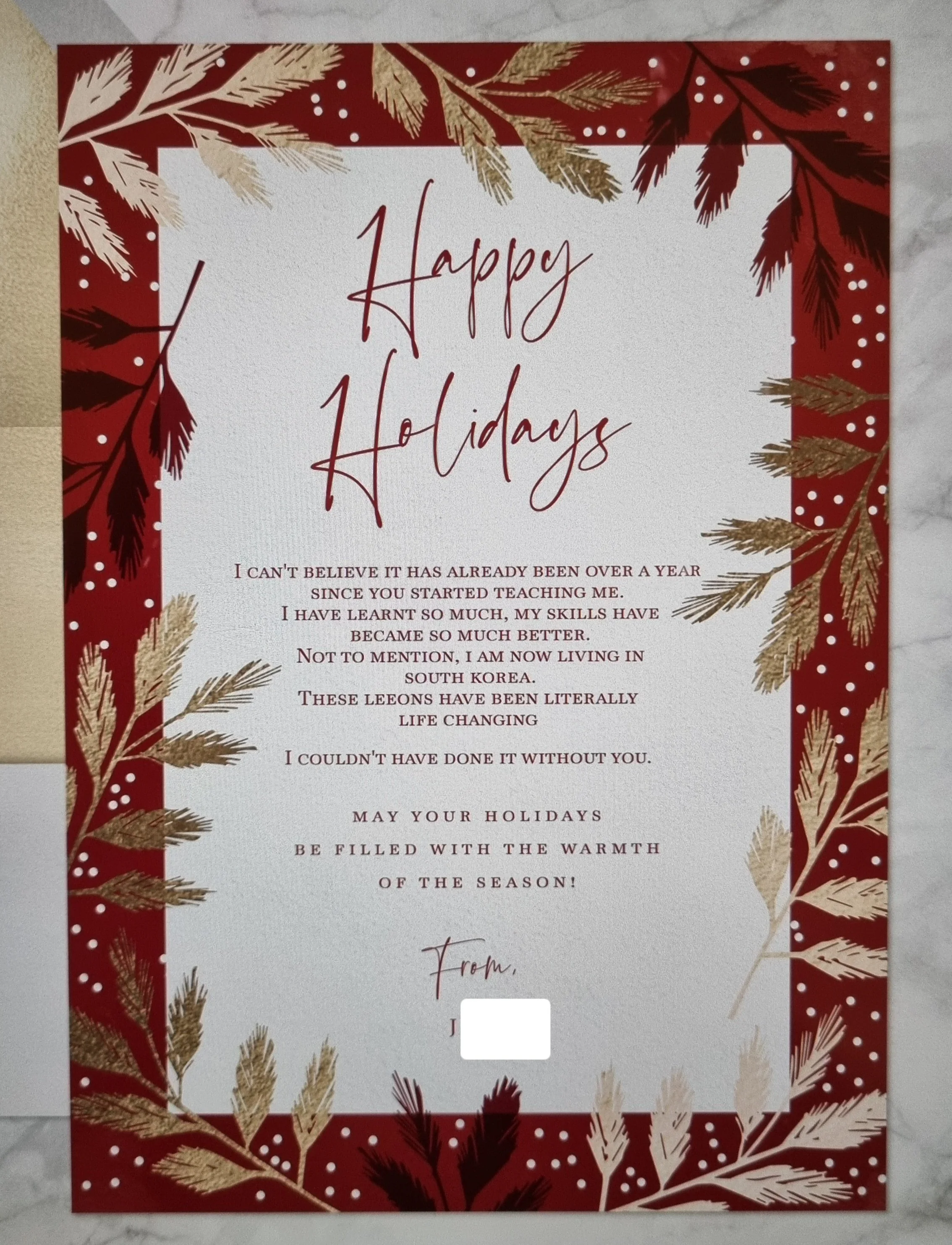 A Christmas greeting card with a red border decorated with gold and dark red pine branches and snow dots. The message inside reads 'Happy Holidays' and contains a personal note about teaching and living in South Korea, ending with holiday wishes.