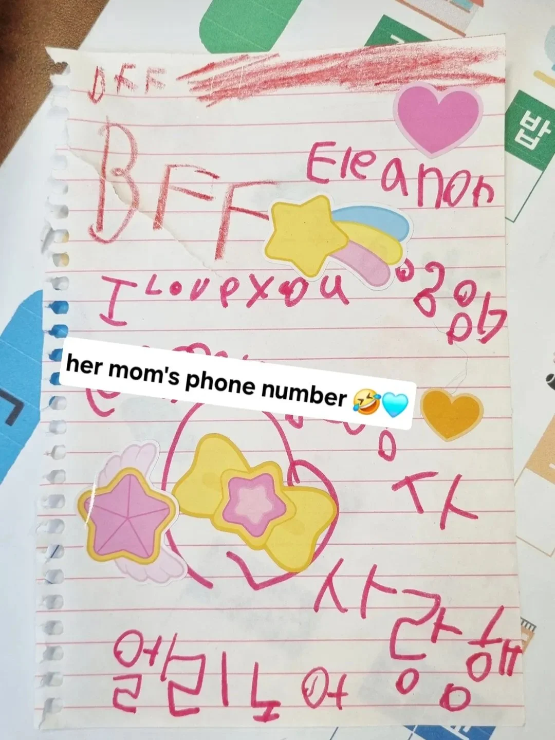 Child's handwritten note on lined notebook paper with colorful star and heart stickers. The note contains handwriting in red and pink, with some words and drawings visible, and a sticker that says "her mom's phone number" with laughing and blue emoji