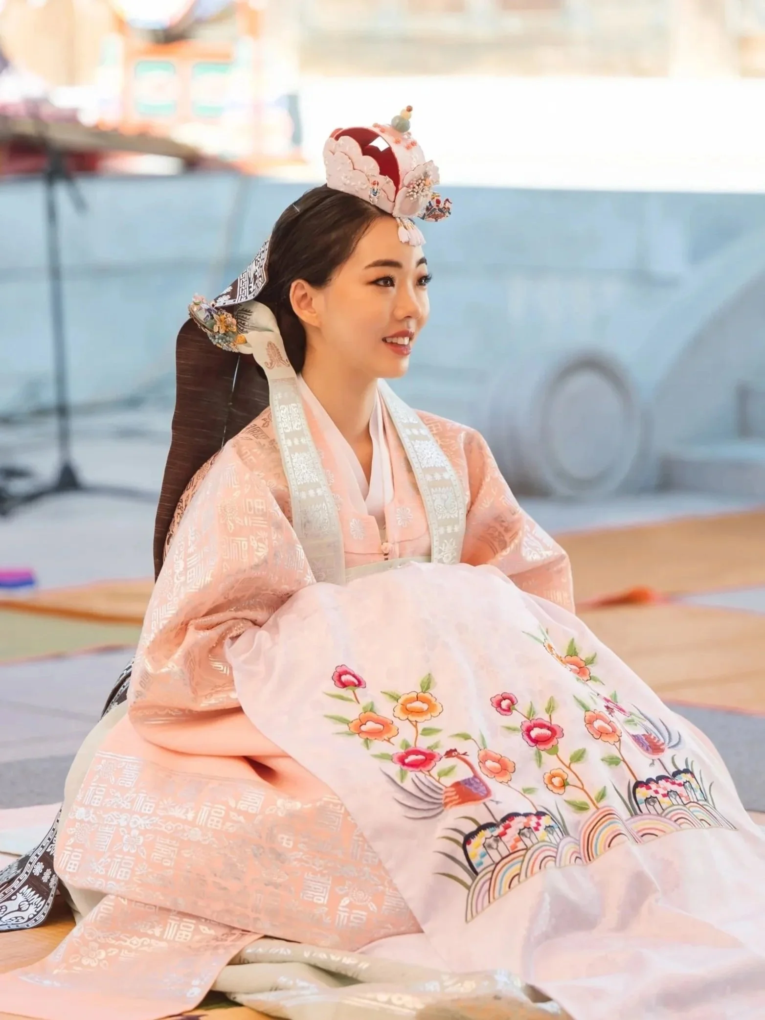 A woman dressed in traditional Korean hanbok sitting at a table, with a decorated headpiece and a soft smile.