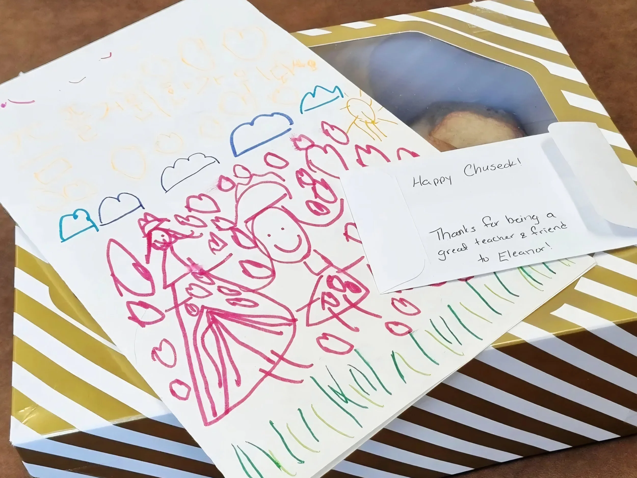Colorful drawing on white paper featuring clouds, hearts, and a smiling figure, alongside a handwritten note saying "Happy Chused!" and a thank you message for a teacher named Eleanor, all placed on a yellow and white striped table.