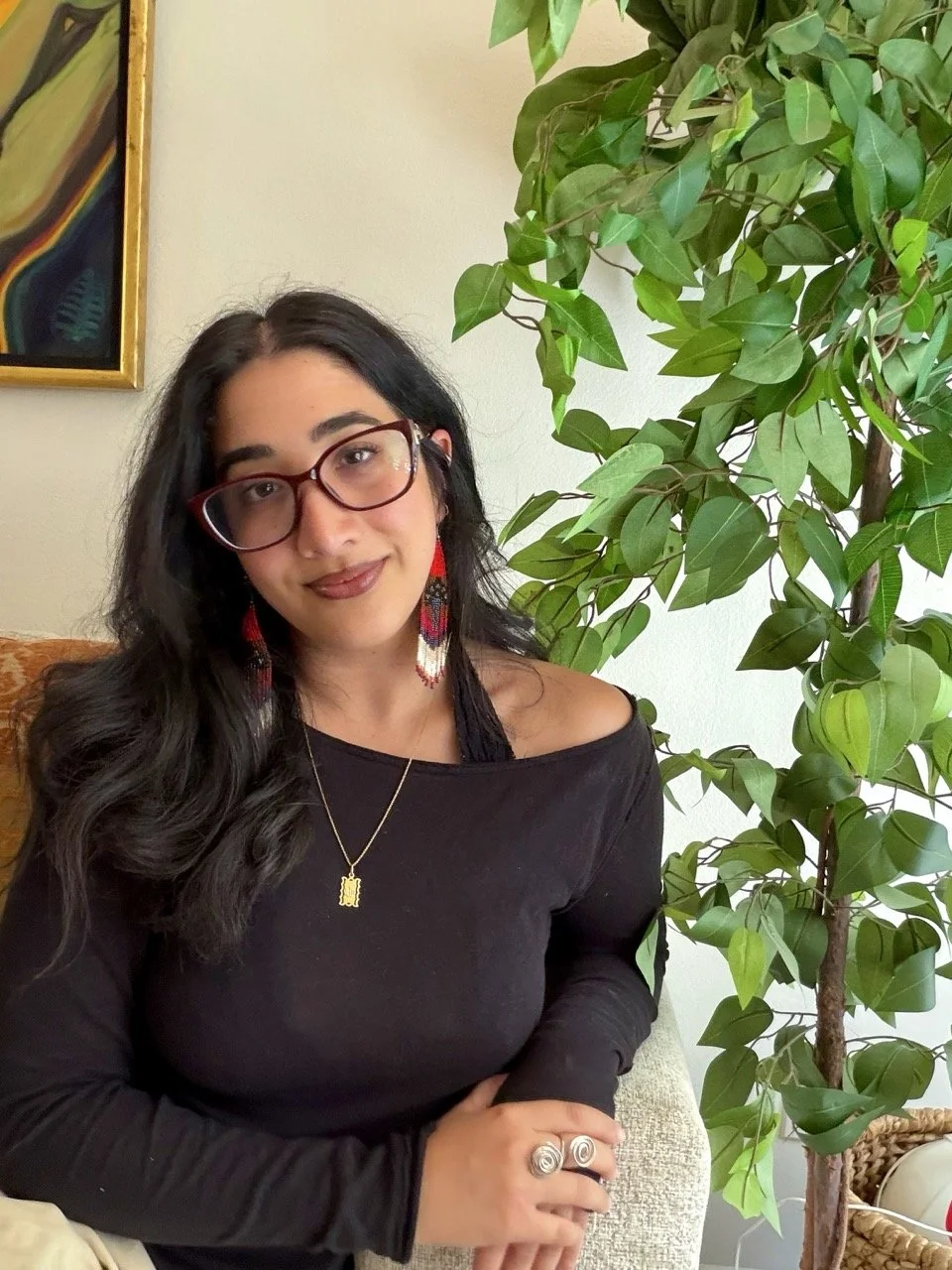 A woman with long black hair, glasses, and earrings, wearing a black off-the-shoulder top, sits beside a large green potted plant indoors.