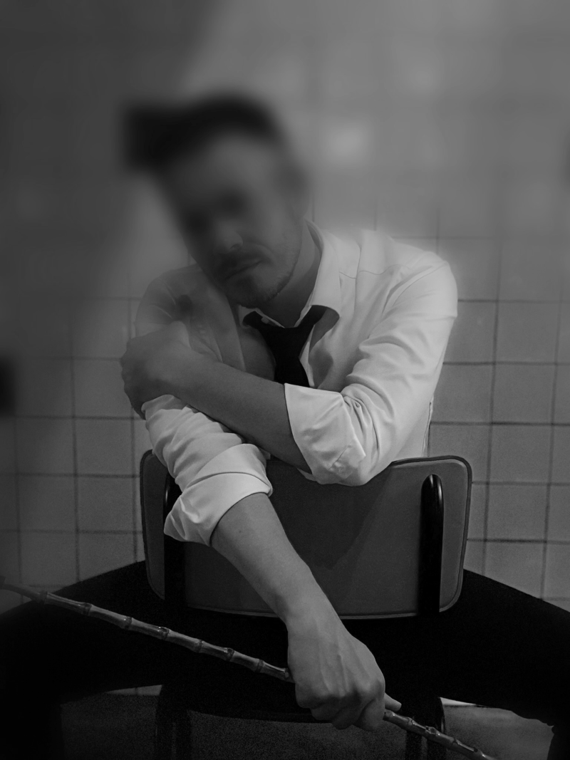 BDSM Gigolo - Een man in een wit shirt en zwarte stropdas zit op een stoel met een muur van tegels op de achtergrond. Hij lijkt te poseren met een stok in één hand.