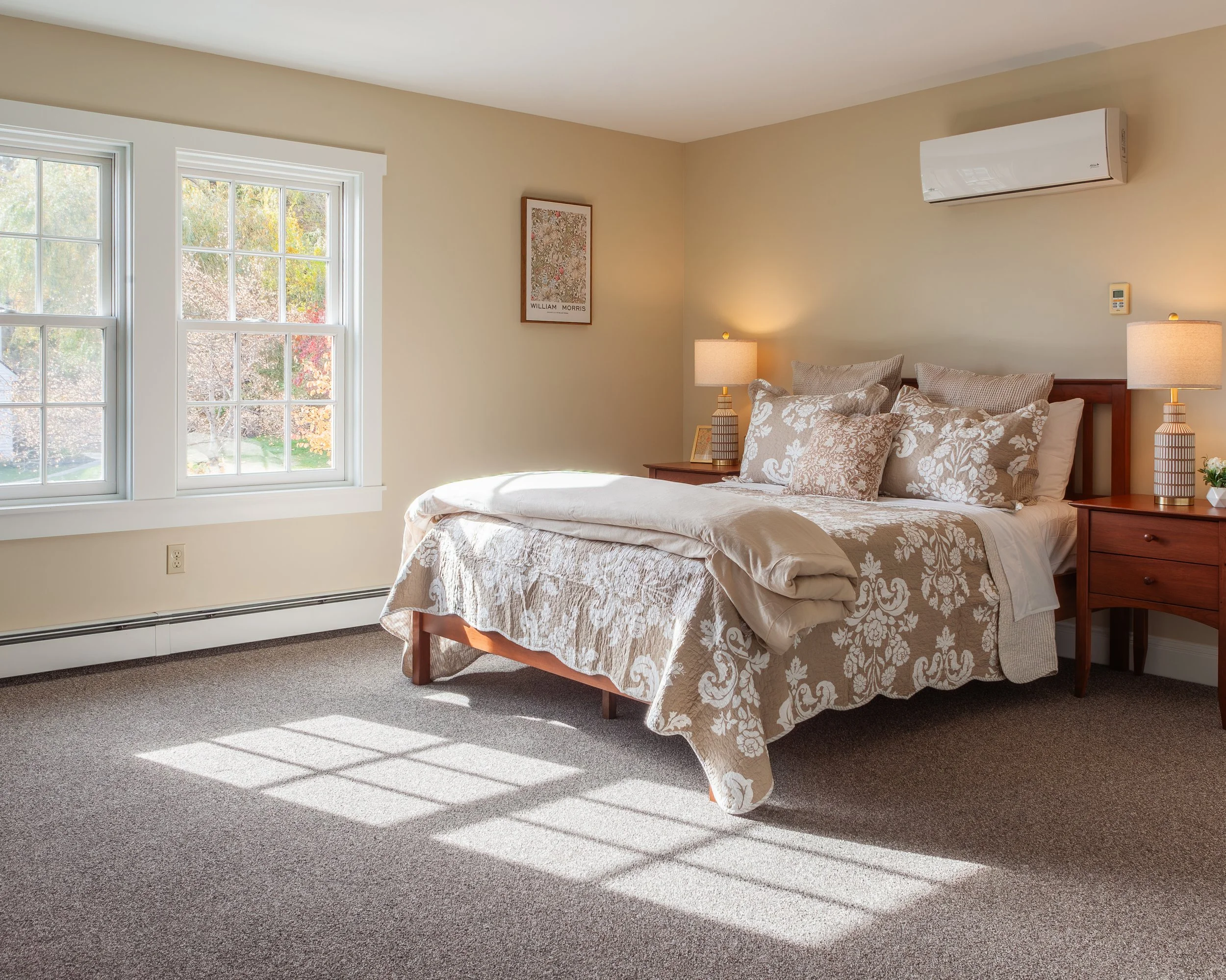 A neatly made bed with beige and white patterned bedding and multiple pillows in a cozy bedroom. There are two bedside tables with lamps, a framed picture on the wall, and sunlight streaming through two large windows with garden views.