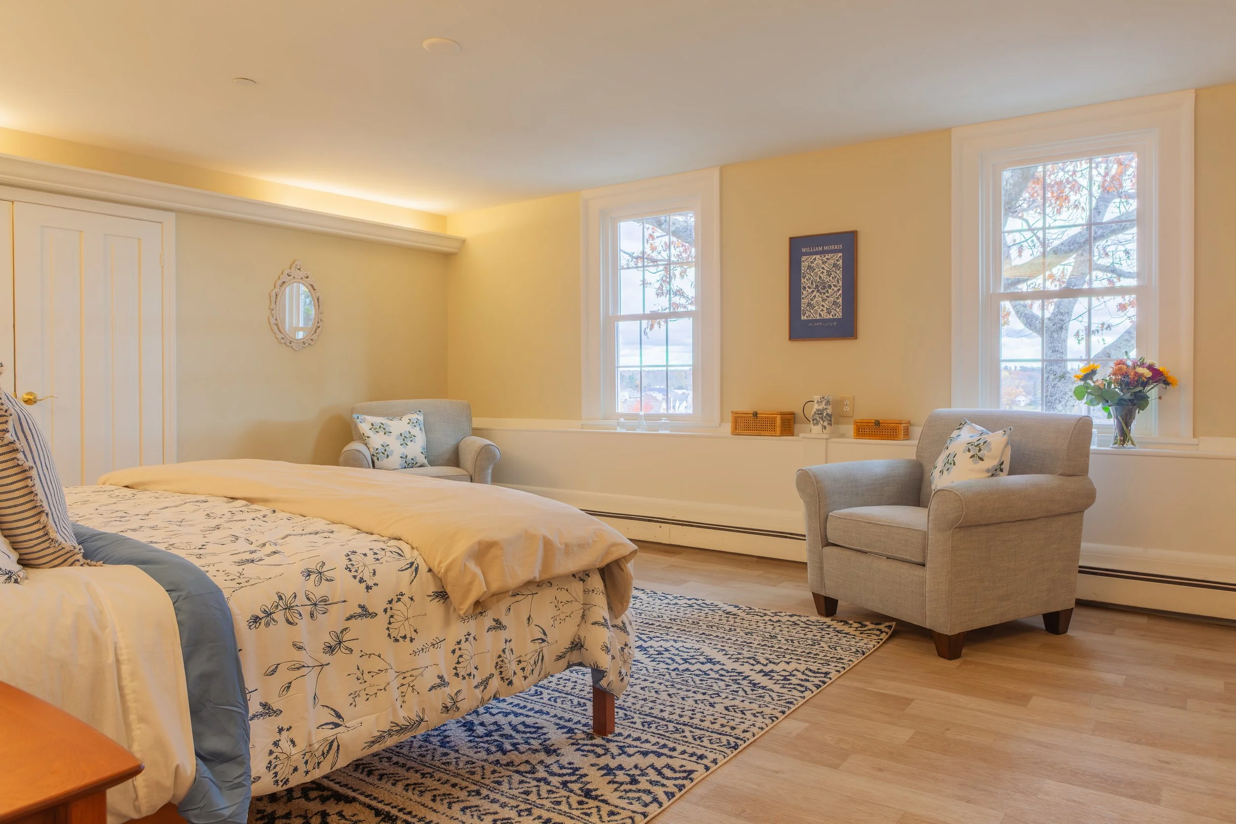 Bright bedroom with two large windows, a bed with floral bedding, and seating area with two armchairs, a rug, and a flower arrangement on the windowsill.