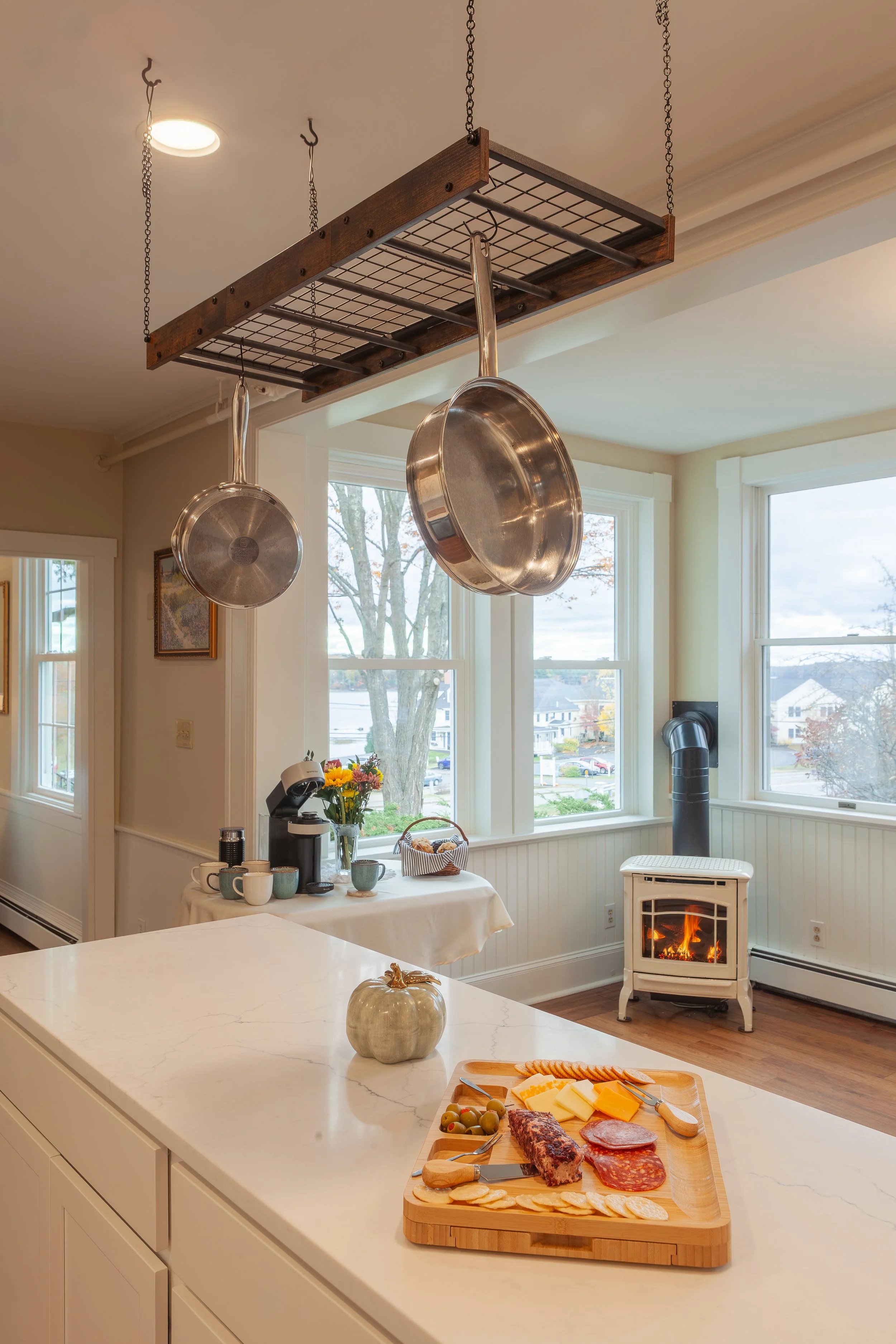 A kitchen with a white countertop, cheese, and sliced meats on a wooden board, a silver pumpkin, a coffee table with cups, a flowers bouquet, and a basket, windows with a view of houses and trees, a small white stove with a fire, and pots hanging fro