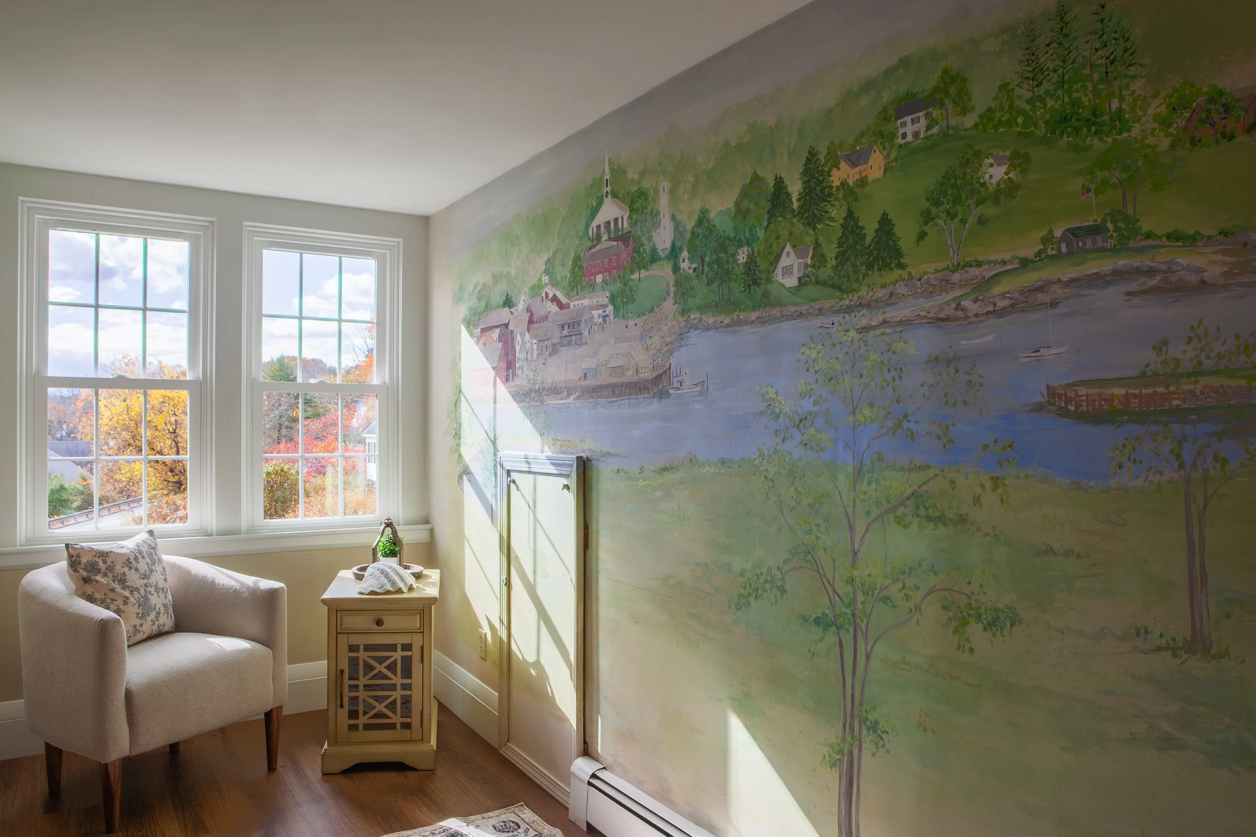 Room with two large windows, a beige armchair with a patterned pillow, a small beige side table, and a mural painting of a lakeside village with houses, trees, and a boat.