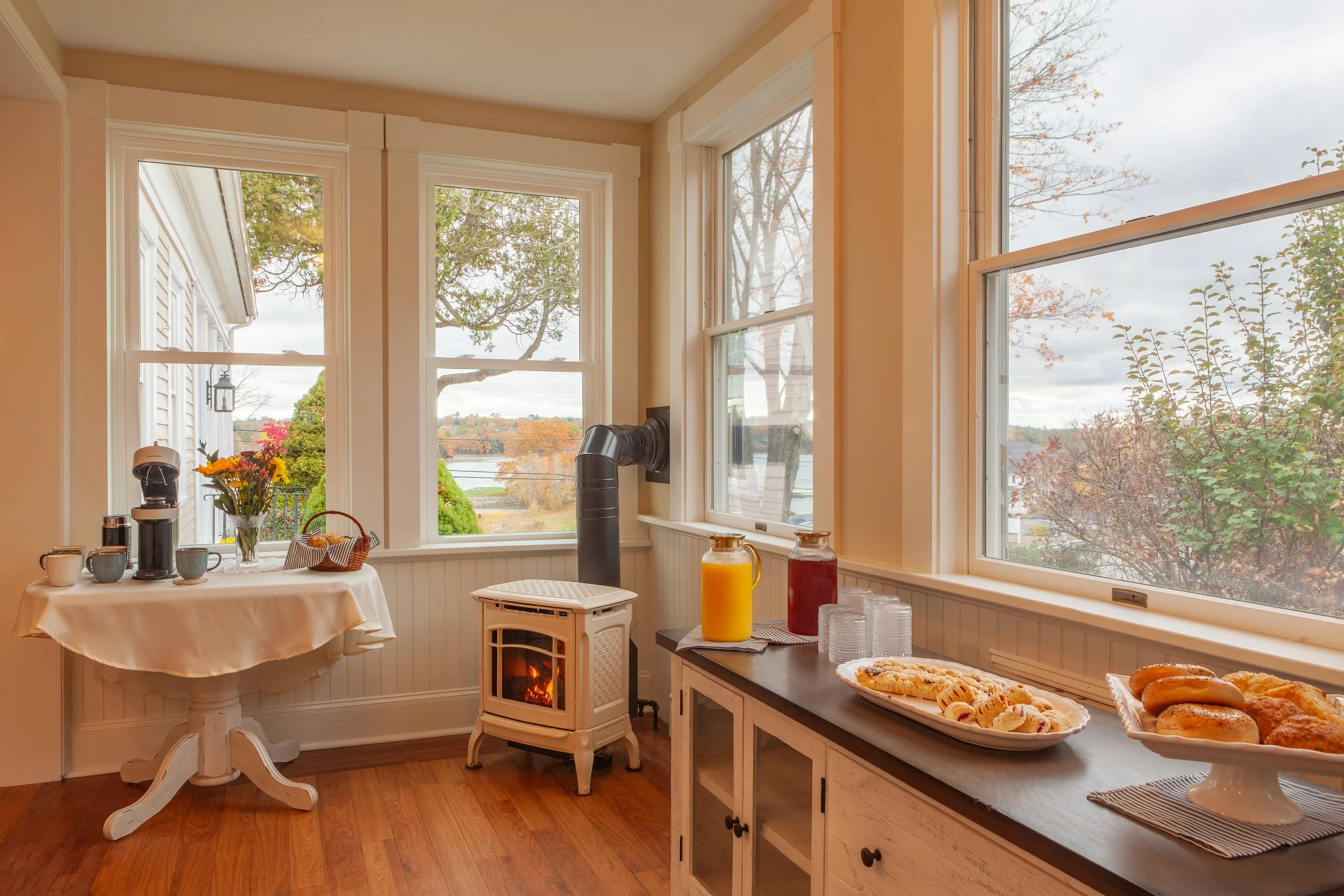 Sunlit breakfast nook with windows, table set with coffee, flowers, and breakfast pastries, and a small stove with a fire.