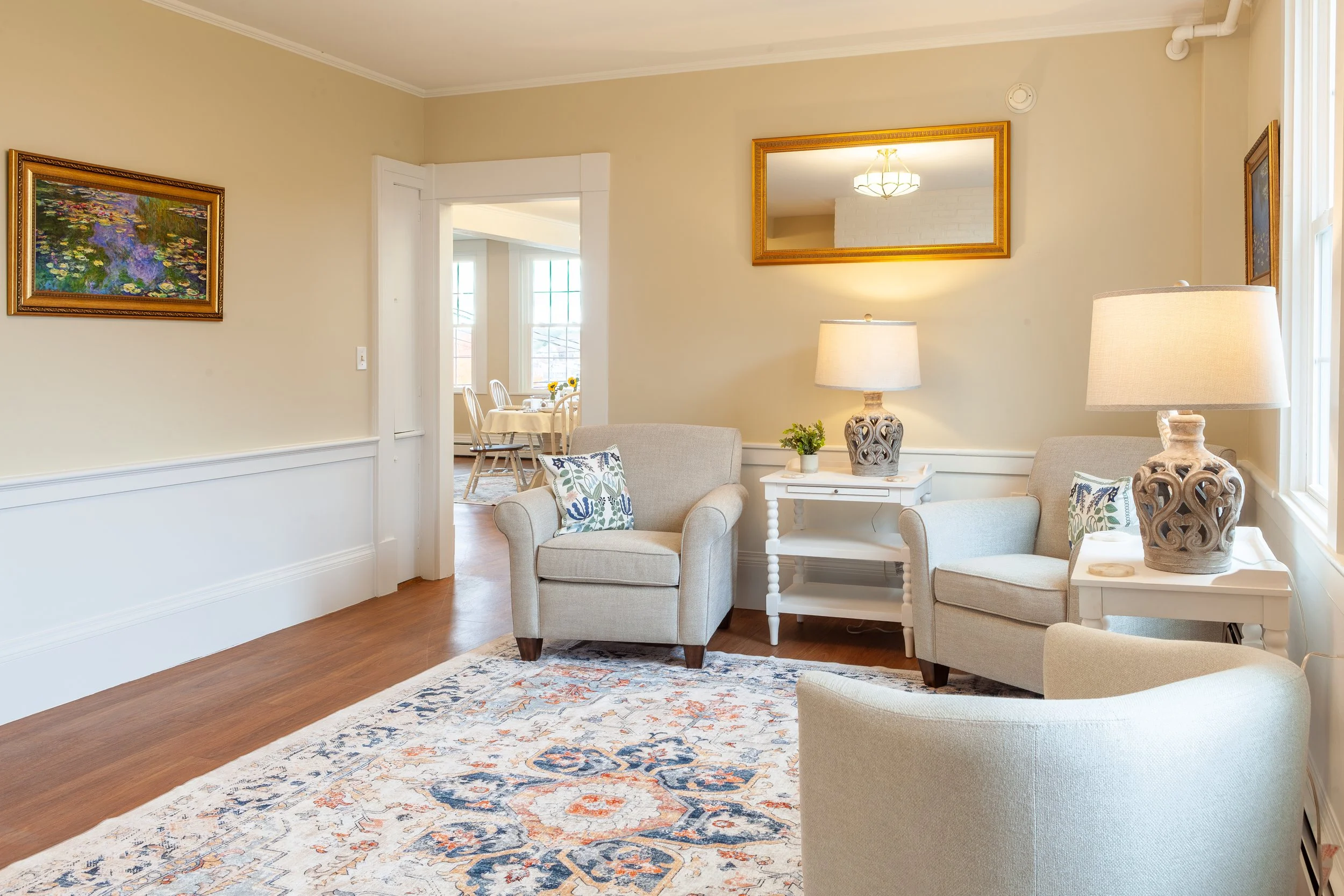 Living room with beige walls, wooden floor, cream armchairs, white side tables with table lamps, framed artwork and a mirror, and a colorful area rug.