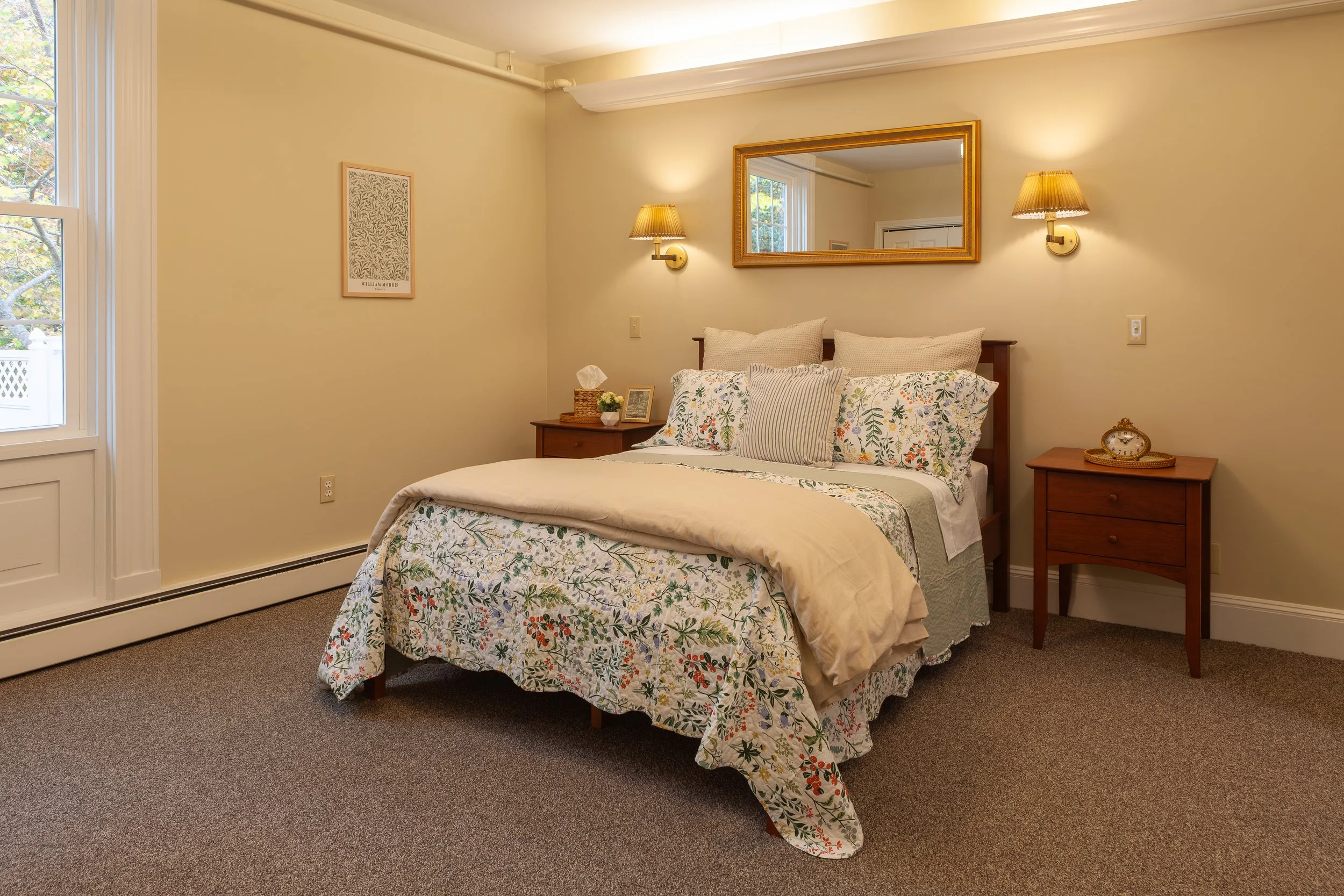 A cozy bedroom with a bed dressed in floral-patterned bedding, wooden nightstands on each side, and a large mirror above the bed. Warm lighting from wall-mounted lamps creates a relaxing atmosphere.