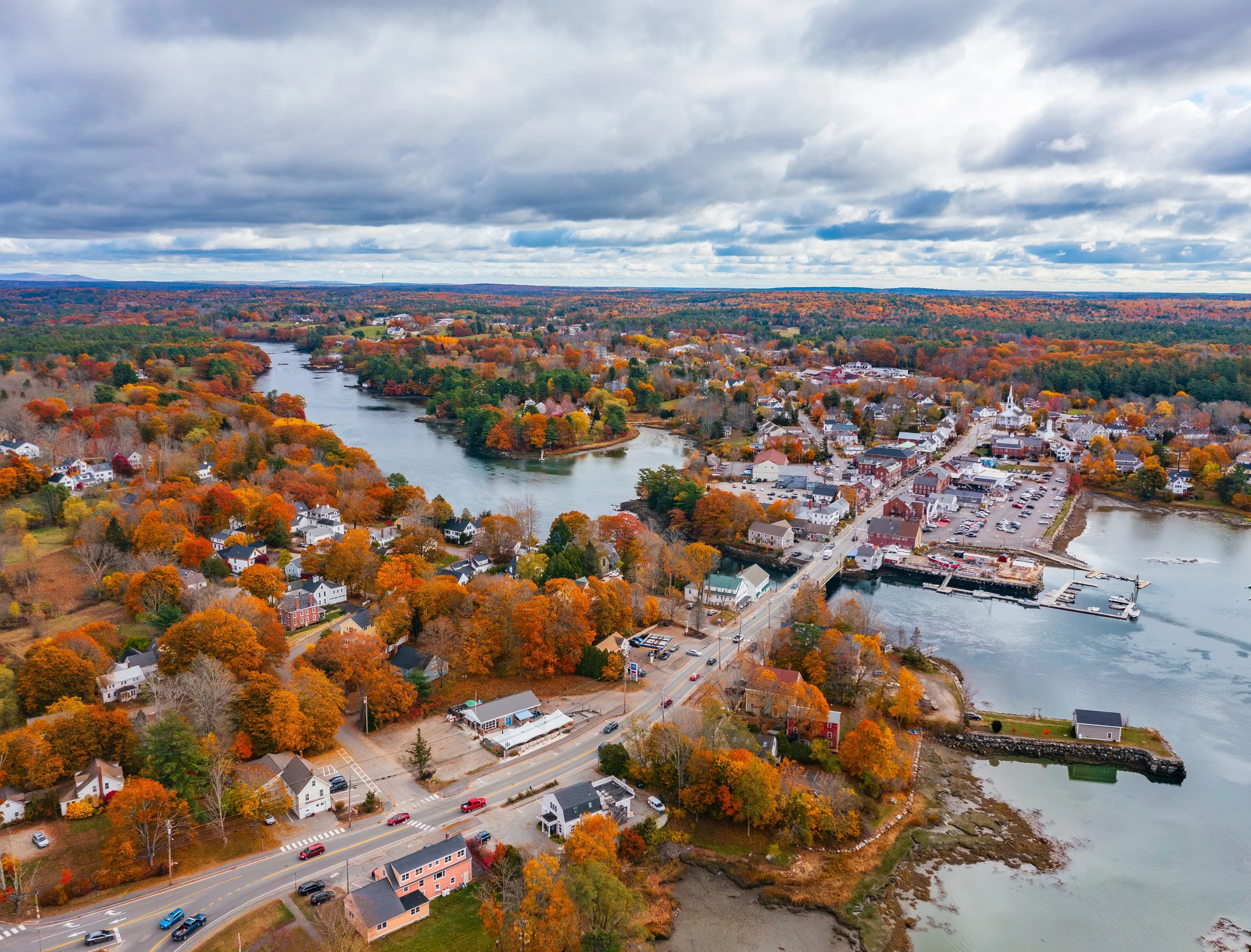 Aerial view of a small town surrounded by water and colorful autumn trees, with a cloudy sky overhead.