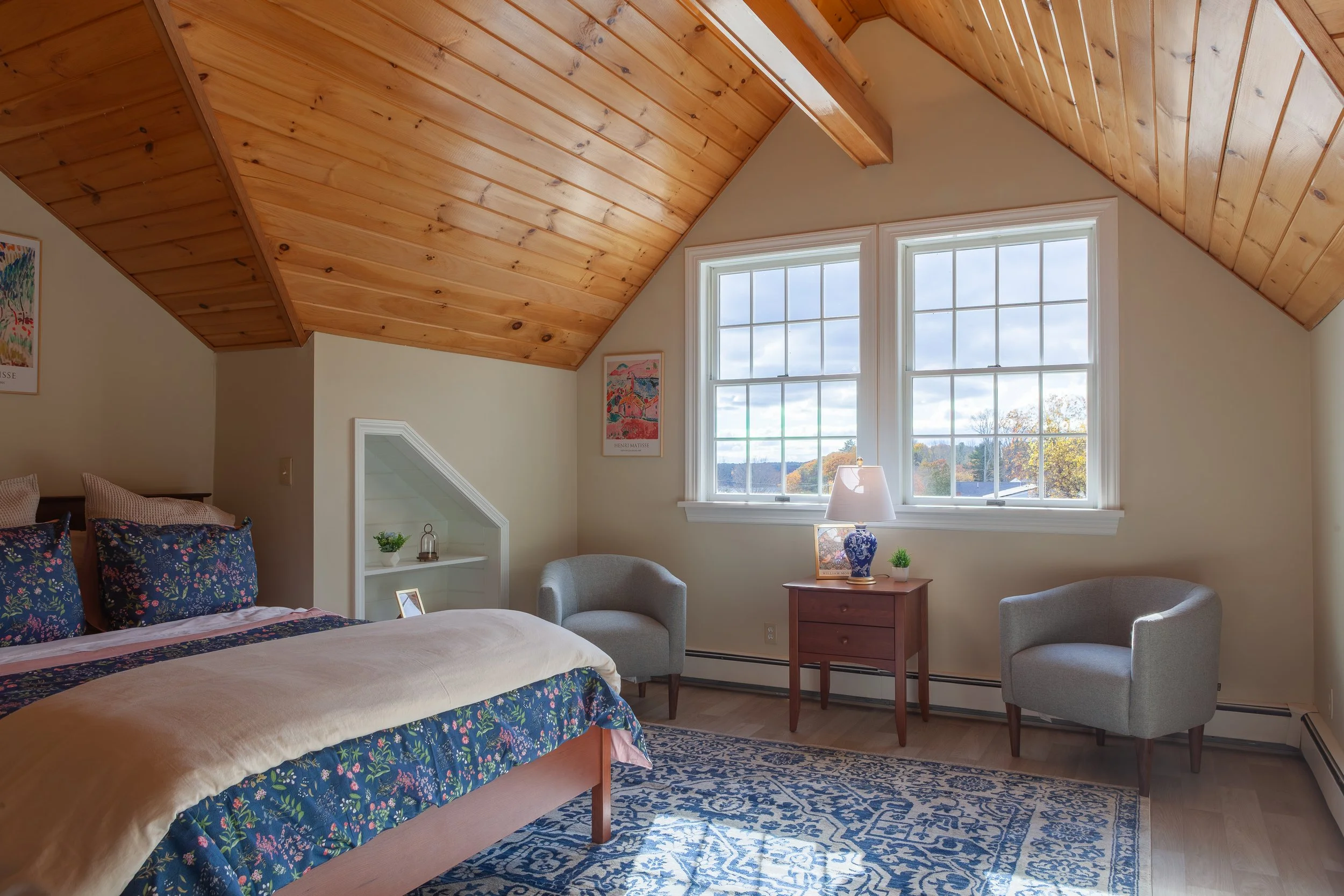 A bedroom with a sloped wooden ceiling, beige walls, and large double windows. There is a bed with floral bedding, two gray armchairs, a small wooden side table with a lamp, and a patterned blue and beige rug.