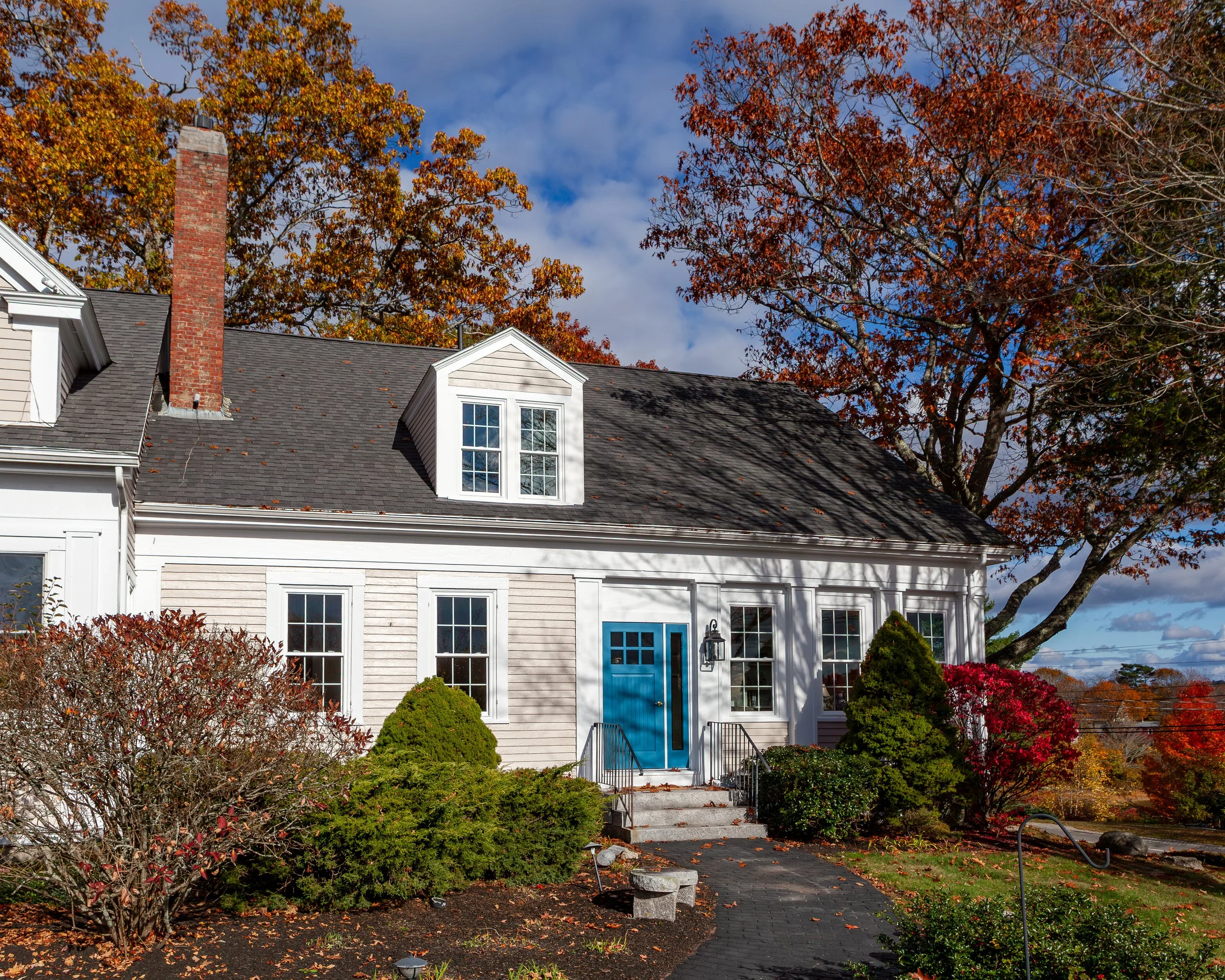 A house with white siding, a blue front door, a brick chimney, and multiple windows, surrounded by trees with autumn foliage.