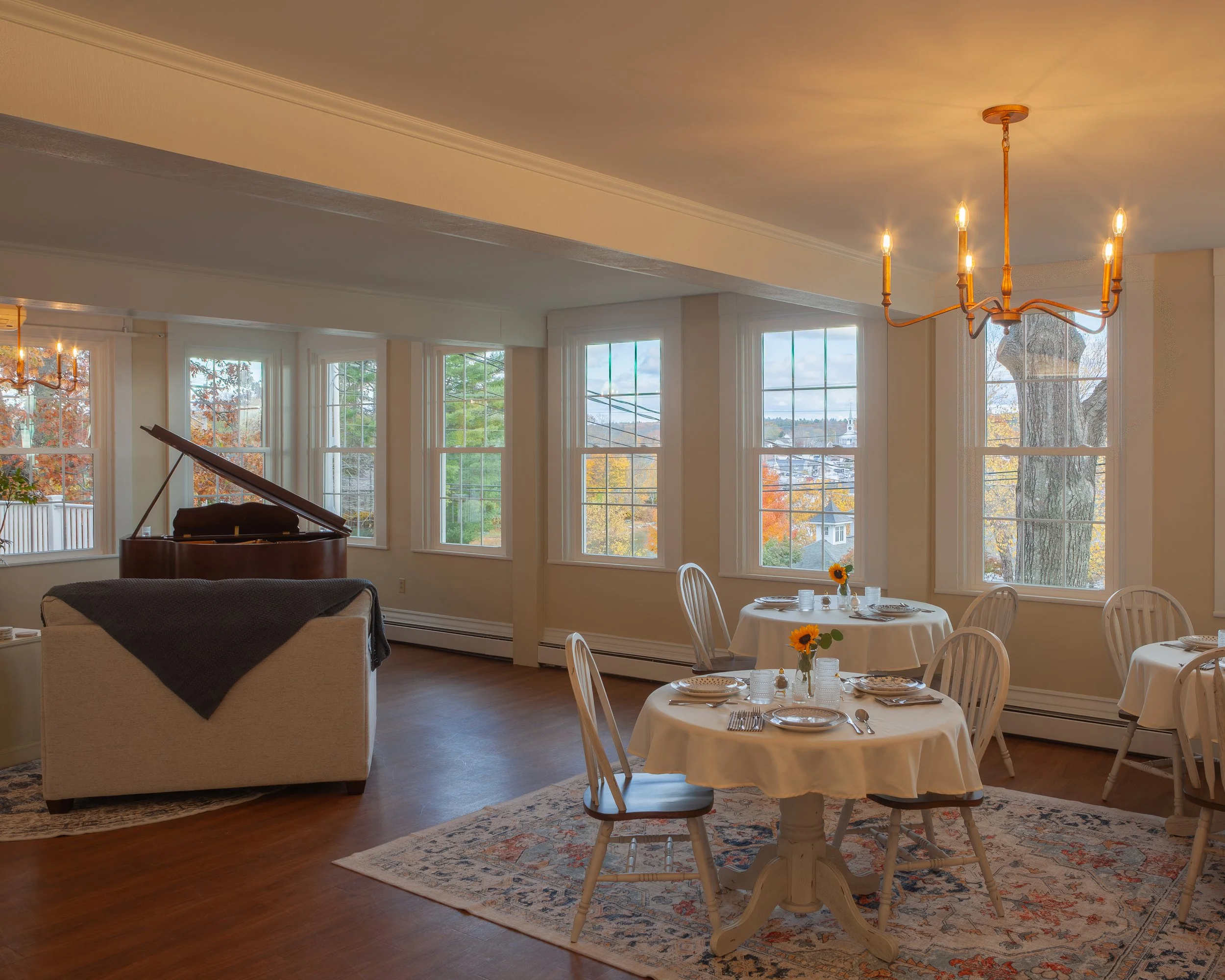 Dining room with round tables set with plates and cutlery, each table decorated with sunflower centerpieces. Large windows with trees and fall foliage outside, hardwood floors, a chandelier overhead, and a grand piano in the background.