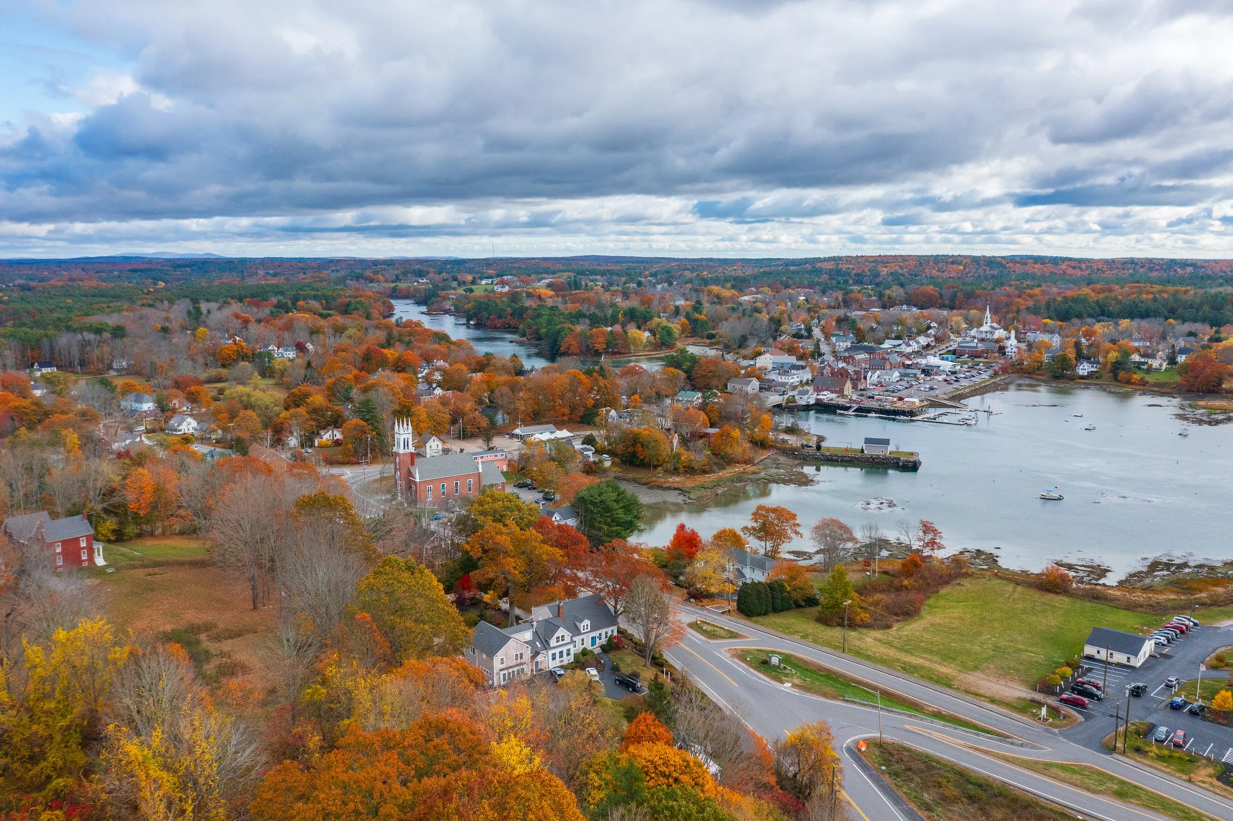 An aerial view of a small town in autumn, with colorful fall foliage, a river running through, and a cloudy sky overhead.