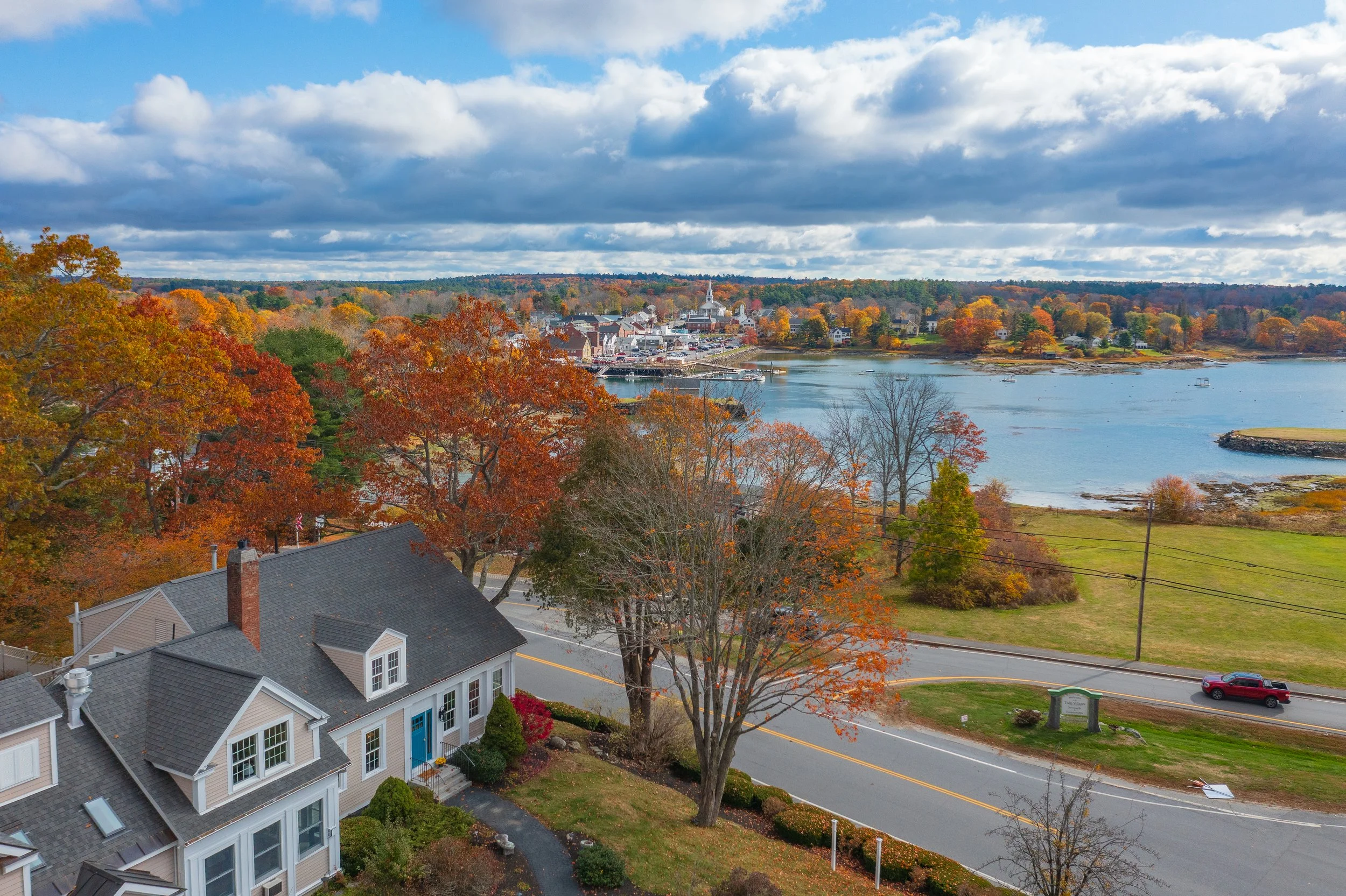 An aerial view of a small town by the water during autumn, with colorful fall foliage and houses near the road.