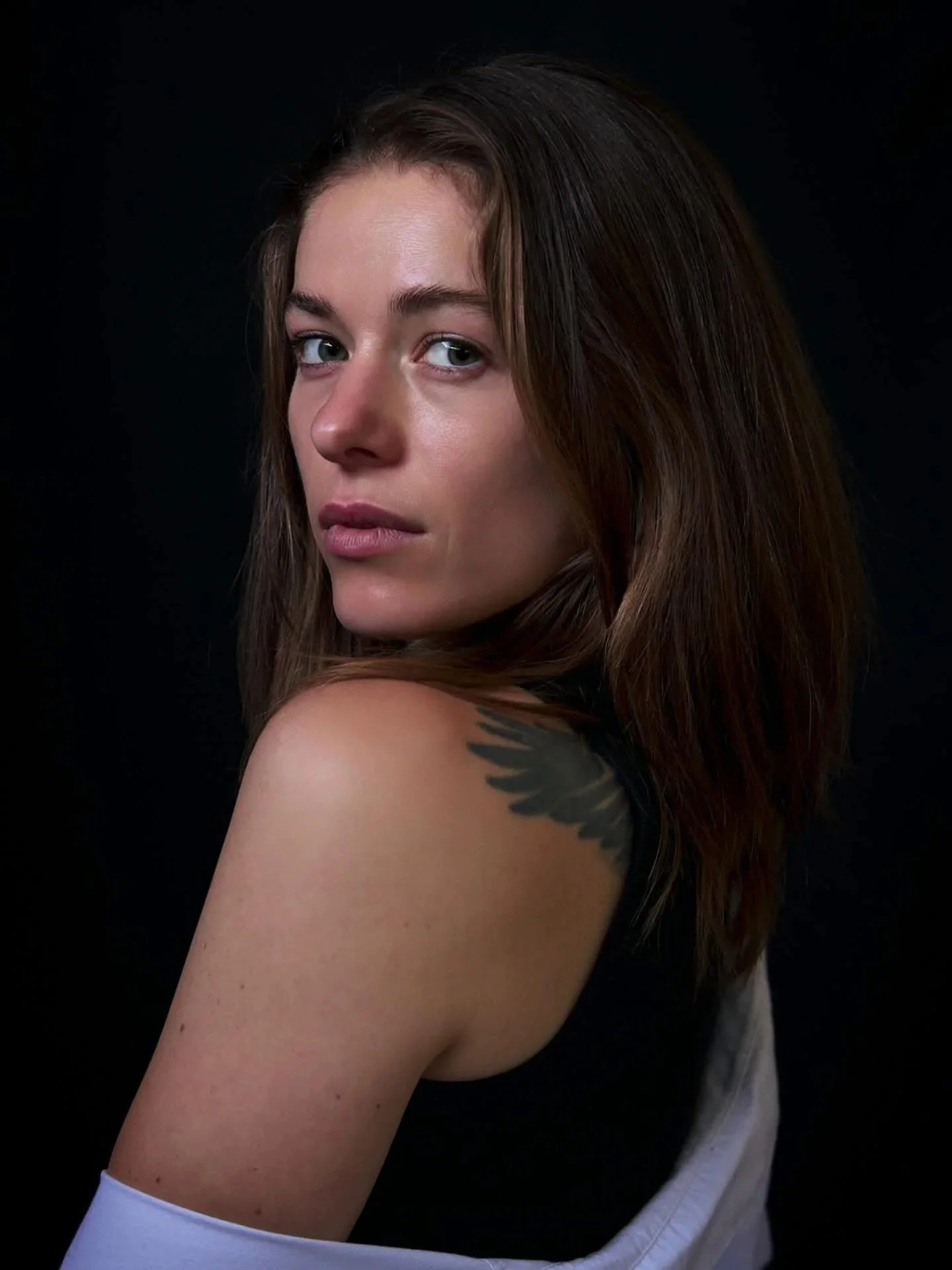 A young woman with long brown hair, looking over her shoulder, with a tattoo of leaves on her upper arm, against a black background.