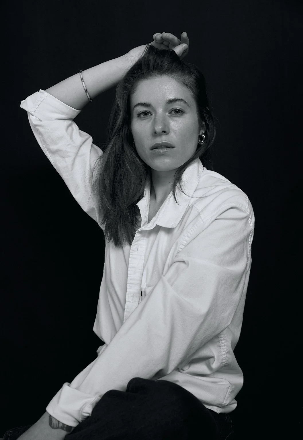 Black and white photograph of a young woman with long hair, one arm raised with hand resting on her head, wearing a white shirt and earrings against a dark background.
