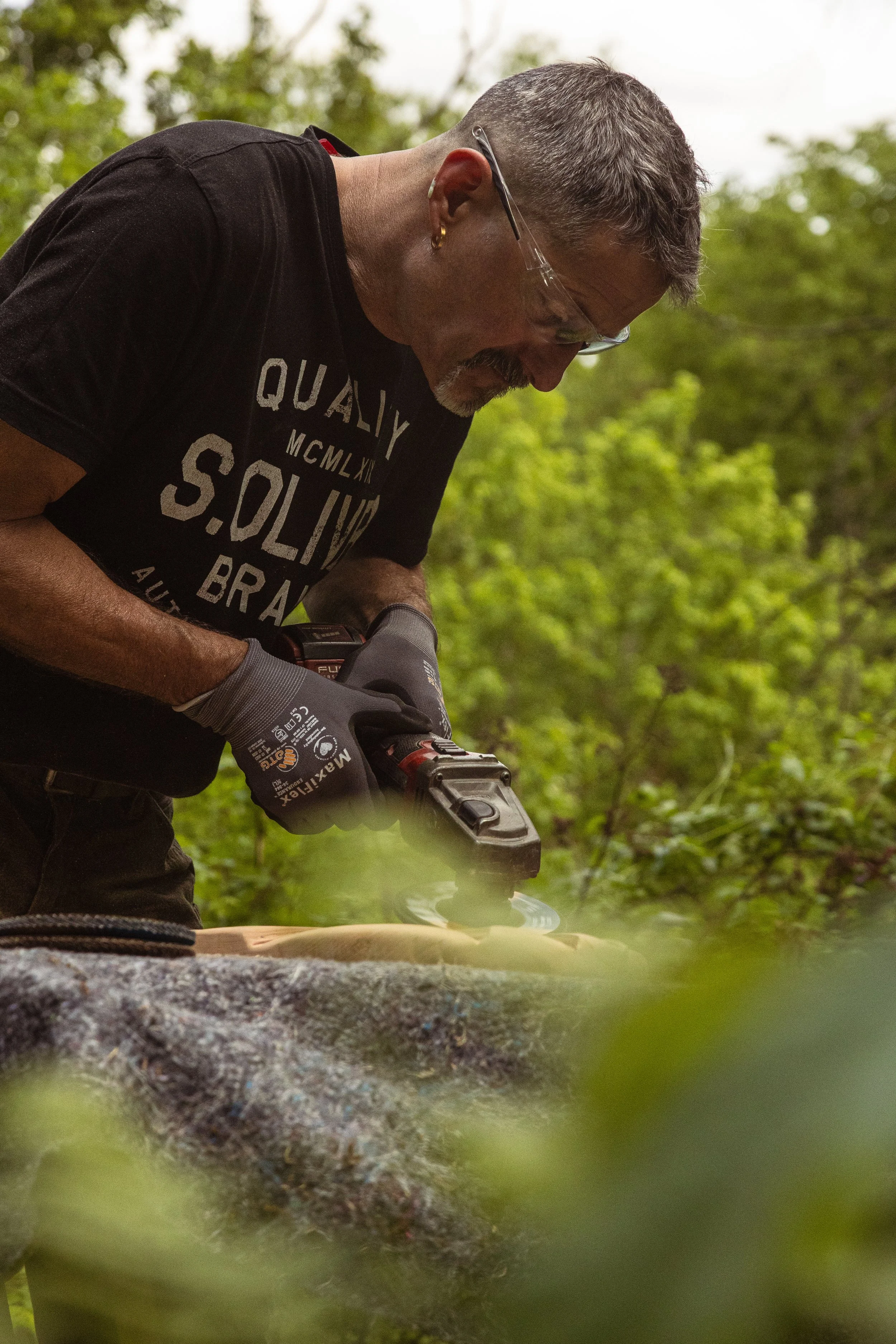 Ein Mann trägt Sicherheitsbrille und Handschuhe, er benutzt ein Elektrowerkzeug, um eine Holz- oder Steinoberfläche zu bearbeiten. Im Hintergrund sind Bäume und grünes Laub sichtbar.