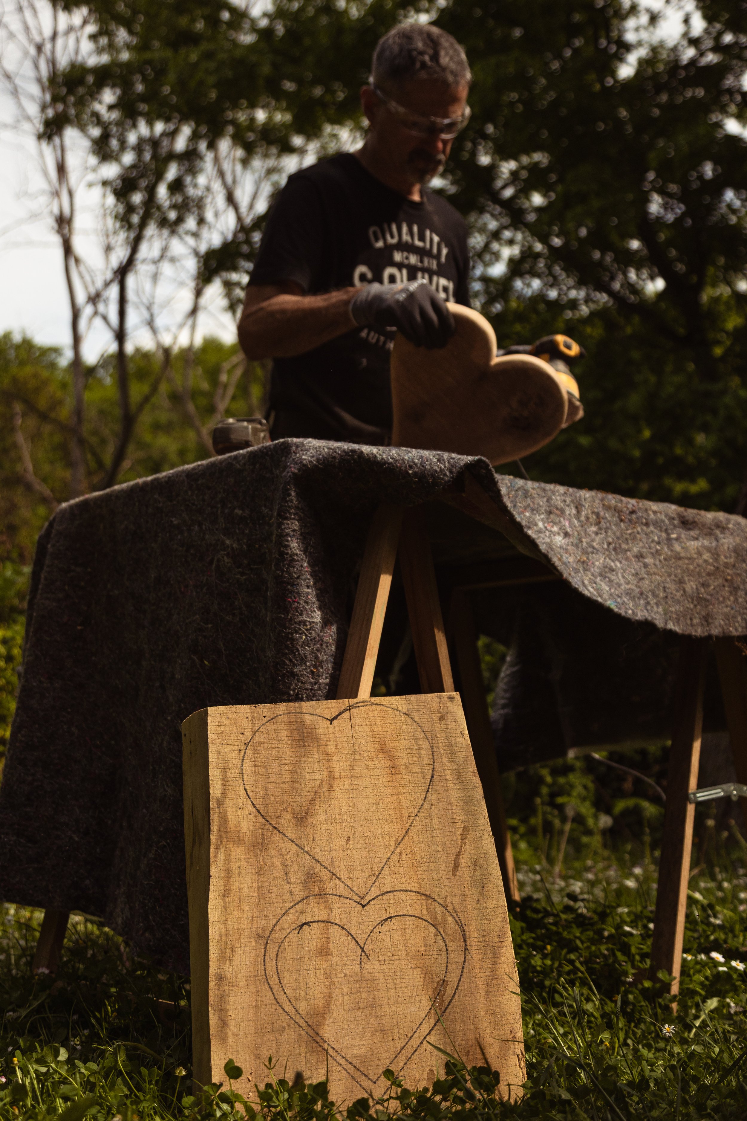 Ein Mann arbeitet im Freien auf einer Holztischliste mit gezeichneten Herzen. Er trägt eine schwarze T-Shirt mit weißen Aufdruck und trägt Schutzbrille, während er ein Holzstück bearbeitet. Im Hintergrund sind Bäume und blauer Himmel zu sehen.