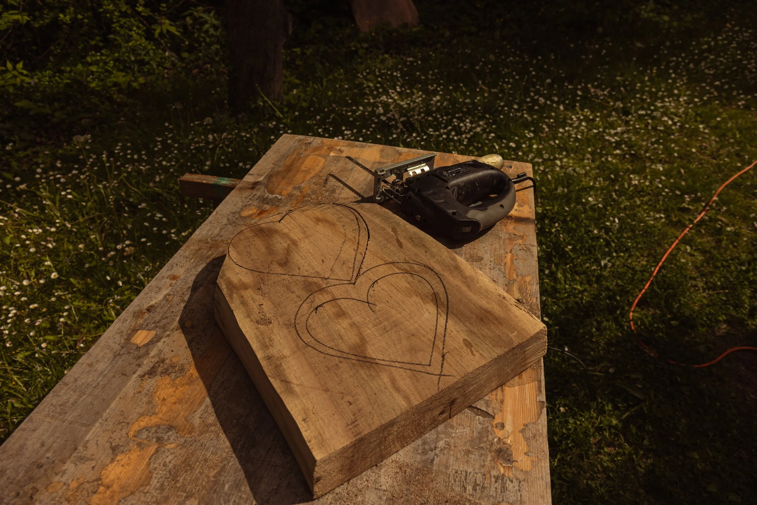 Holzstück mit Kreismustern, elektrisches Sägen auf einem Wackeltisch, draußen in der Dunkelheit, mit Blumen im Hintergrund.