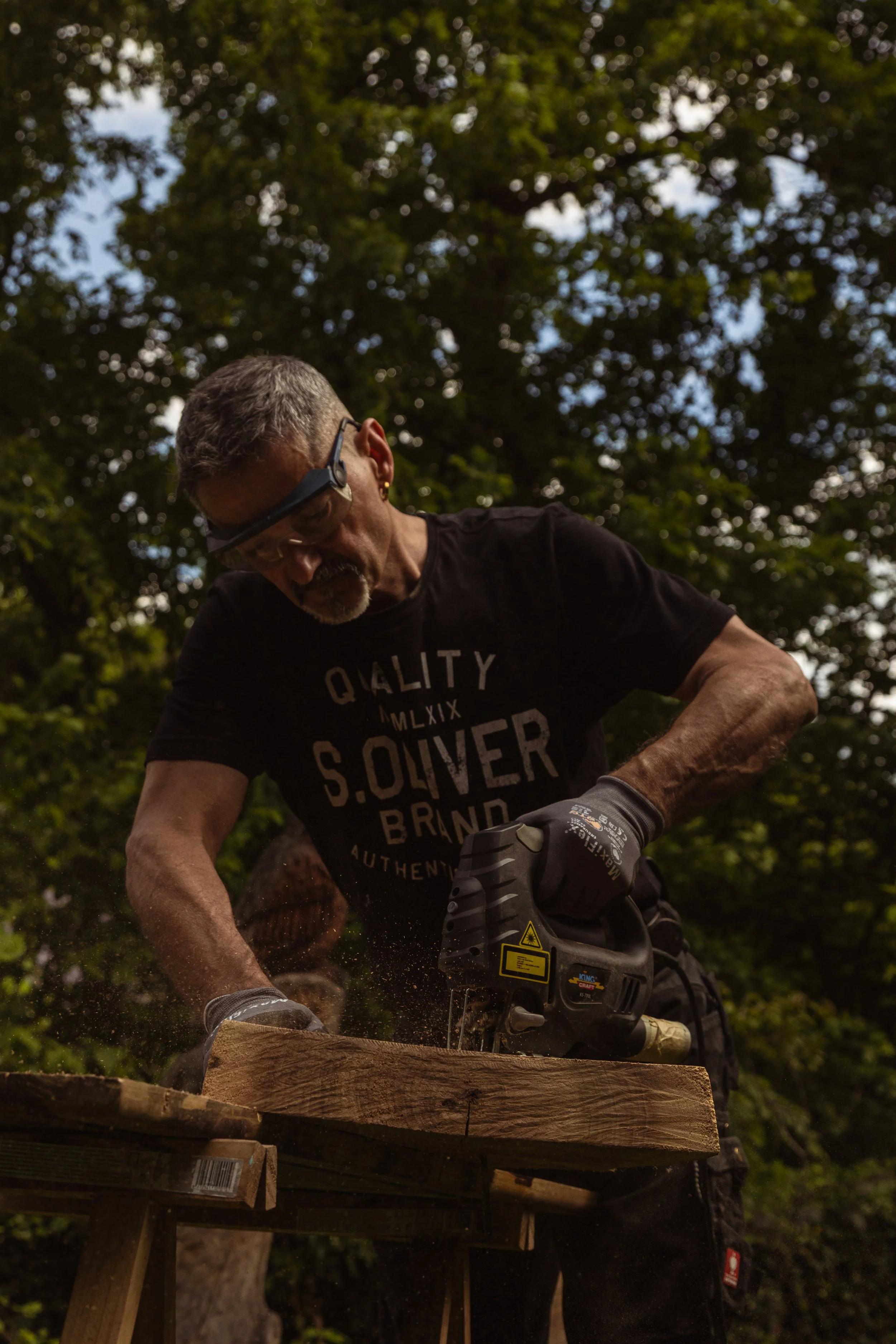 Ein älterer Mann beim Sägen eines Holzstücks im Freien, getragen von Bäumen, trägt schwarze Kleidung und Arbeitshandschuhe, trägt eine Schutzbrille und einen Gehörschutz.