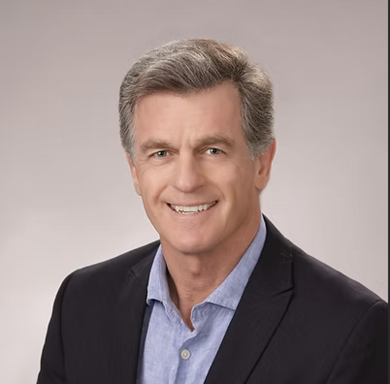 A middle-aged man with gray hair, wearing a black blazer and a light blue shirt, smiling against a neutral background.