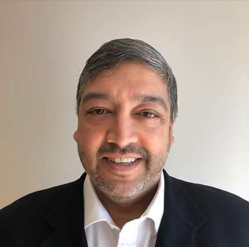 A man with gray hair and beard, wearing a black suit and white shirt, smiling in front of a plain background.